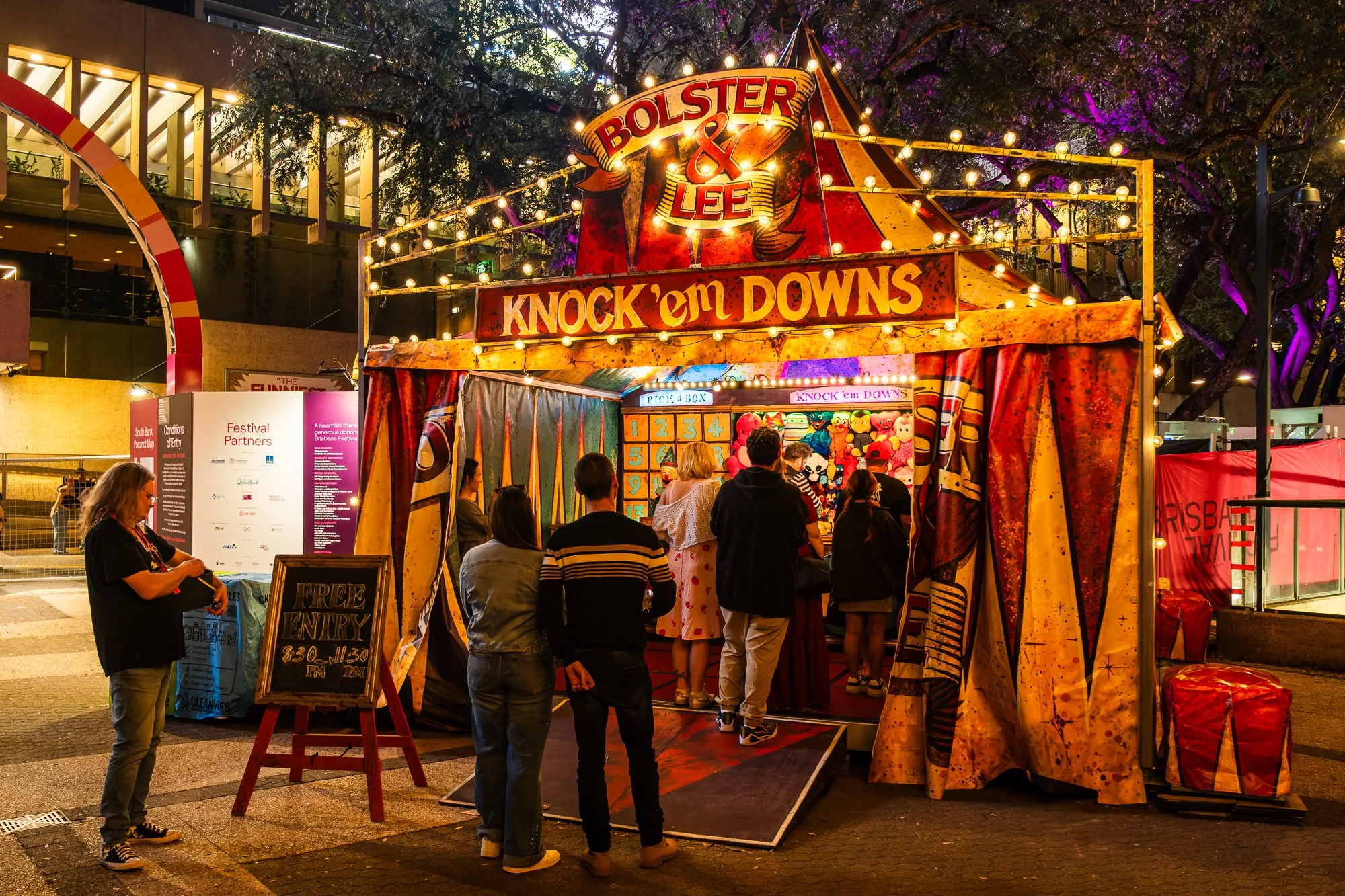 Bright carnival game booth with a sign reading 'Bolster & Lee Knock 'em Downs' illuminated with string lights at night. Several people are lined up in front, playing games, with plush toys as prizes. There is a woman standing next to a sign that says