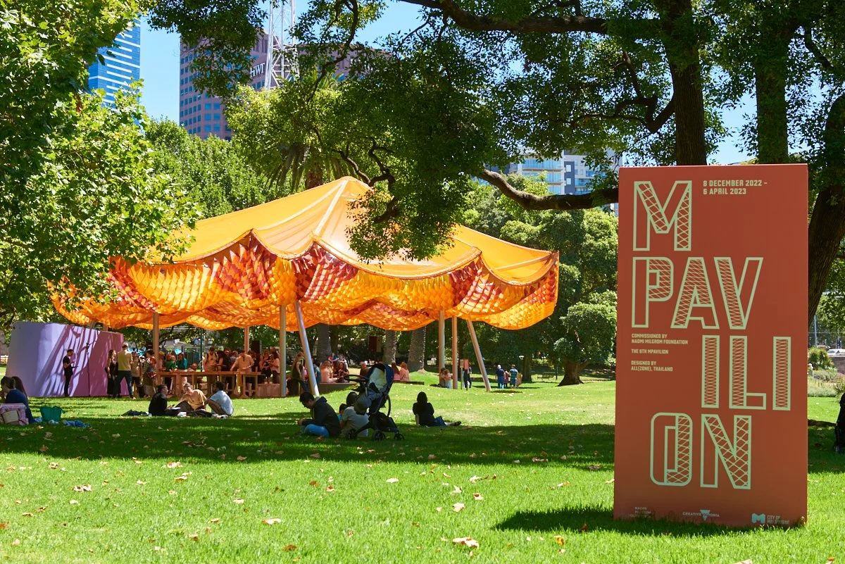 People are gathered in a park under a large yellow canopy tent with orange accents, surrounded by green trees and grass, with tall buildings in the background. A signboard in the foreground reads 'IM PAVILION' with event dates from December 6, 2022, 