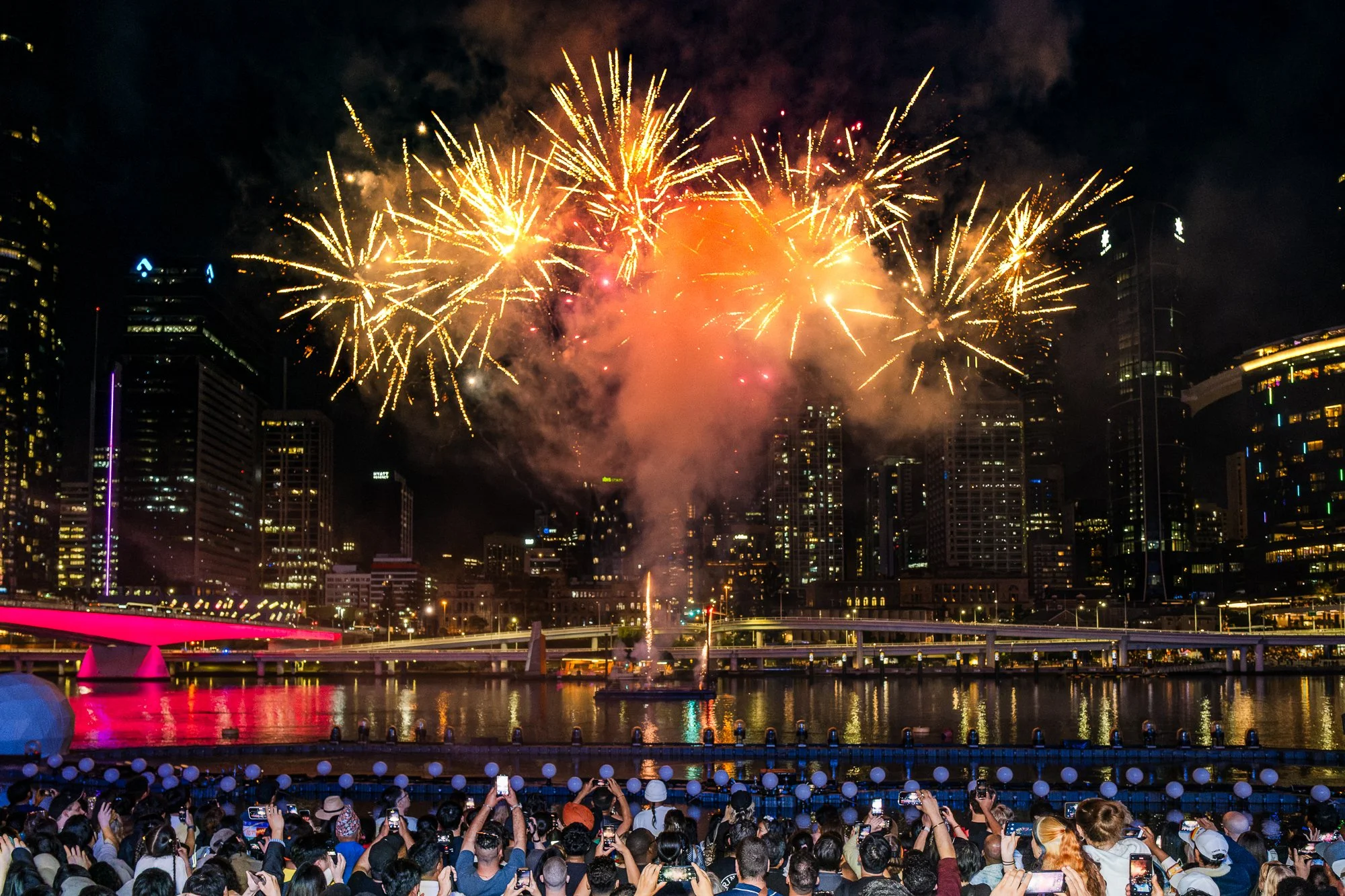 Nighttime cityscape with fireworks over a river, multiple spectators taking photos, and illuminated buildings in the background.