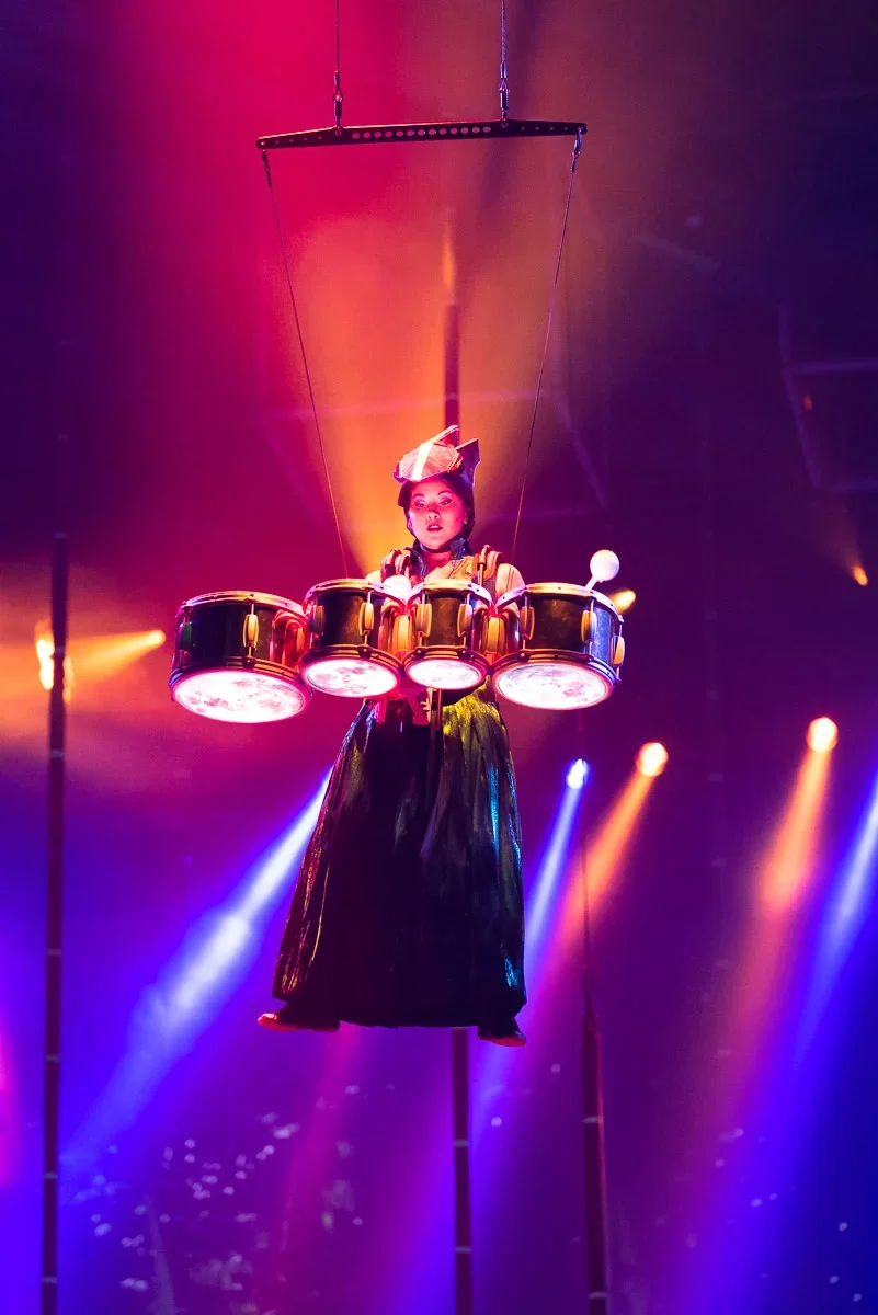 Performer wearing an outfit resembling a drum kit, suspended in the air on a harness, under colorful stage lighting with purple, orange, and blue hues.