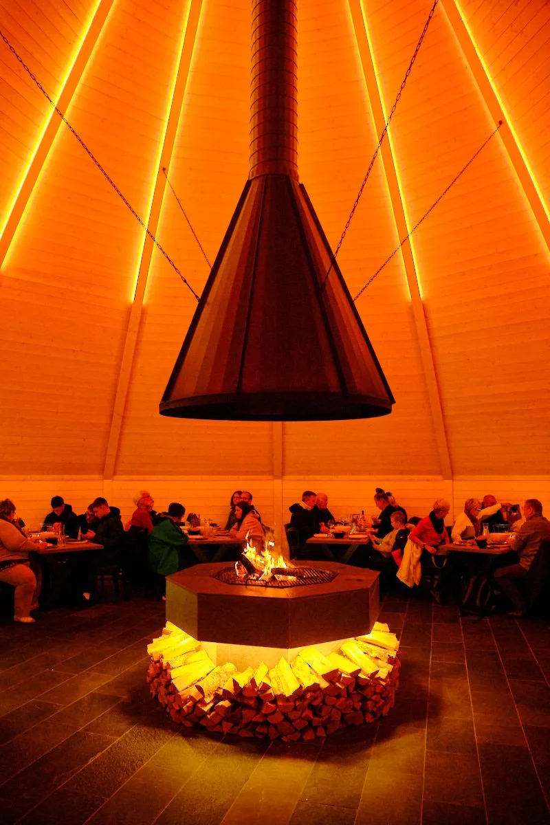 People dining in a cozy restaurant with a central fireplace and a large hanging copper chimney, illuminated by warm orange lighting.
