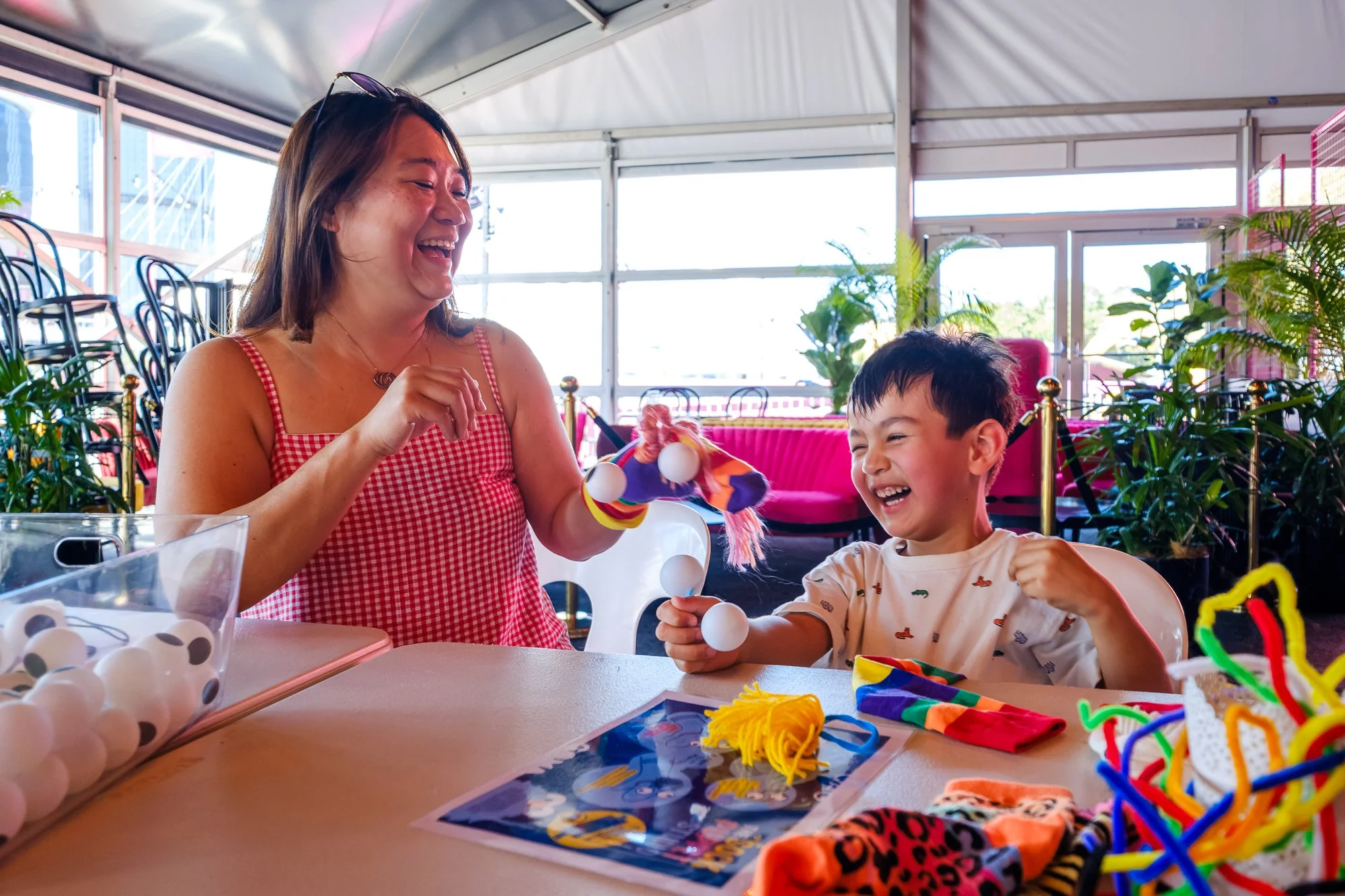 A woman and a young boy playing with a string of white plastic balls at a table, with a pink chair and green plants in the background.