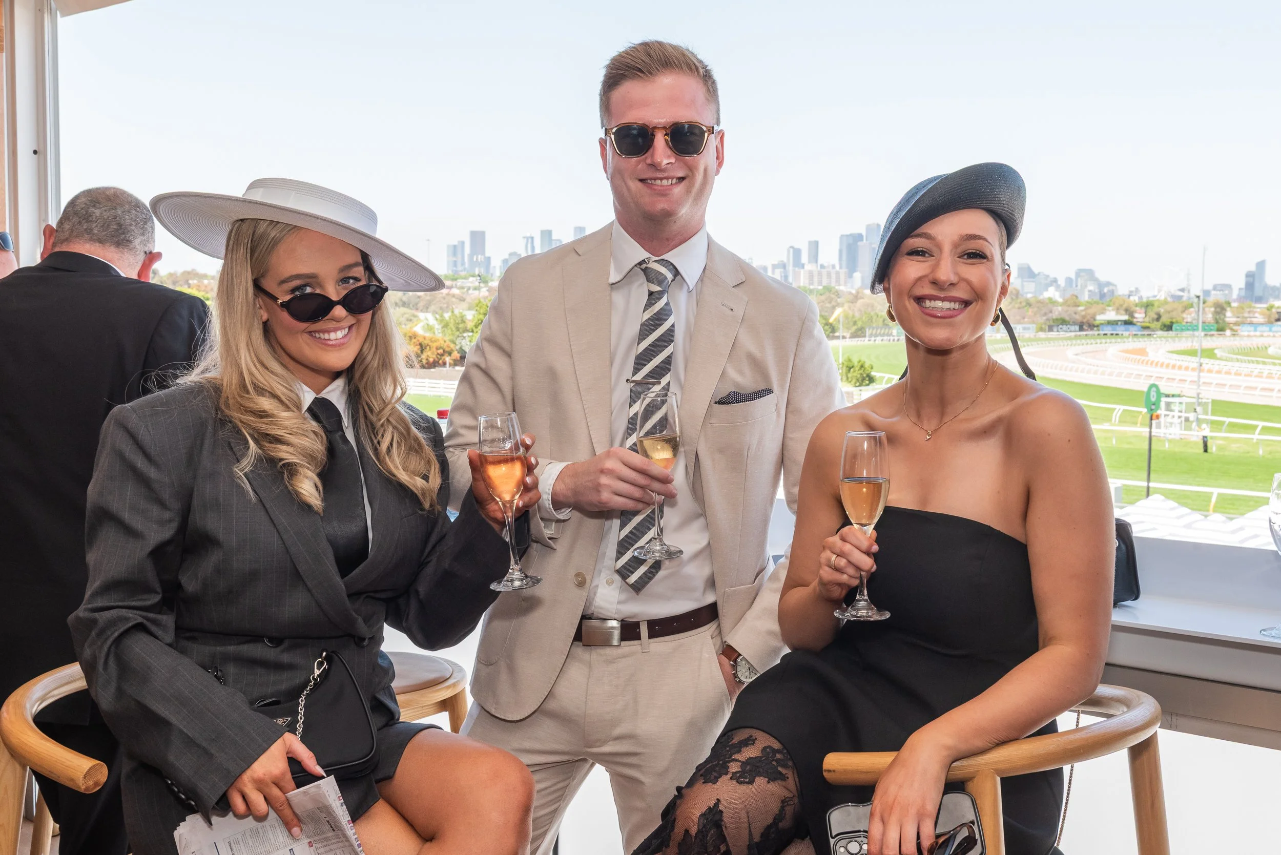 Three elegantly dressed people holding glasses of champagne at a daytime outdoor event with a racetrack and city skyline in the background.