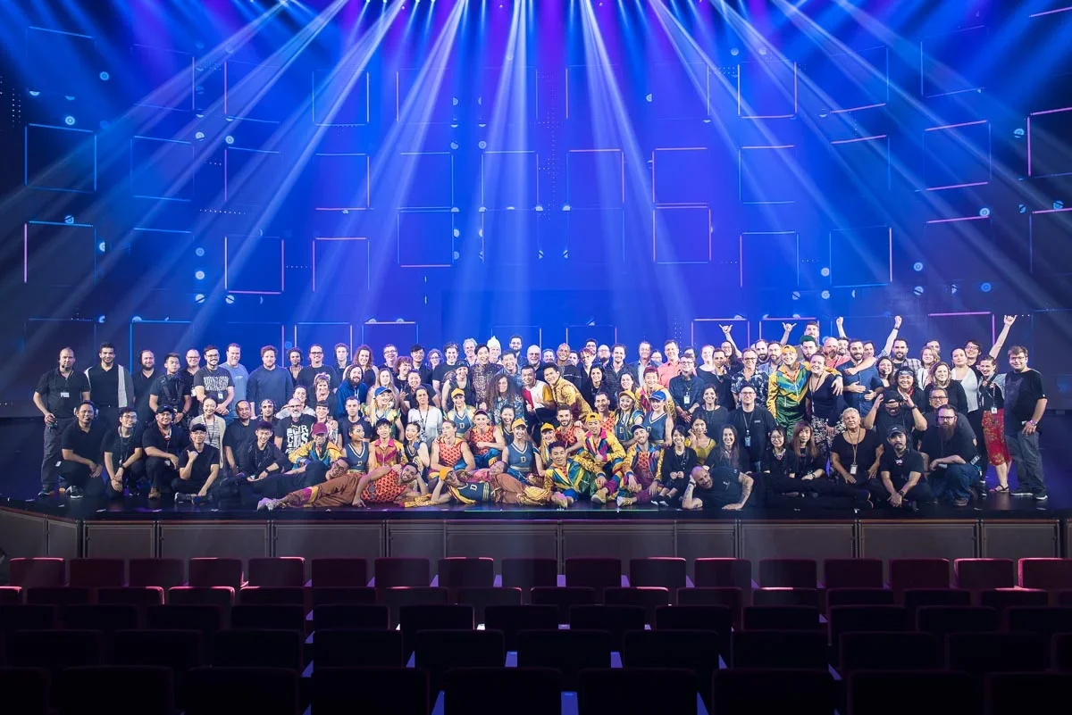 A large group of people on a stage under colorful lights in a concert hall, posing for a group photo, with empty theater seats in the foreground.