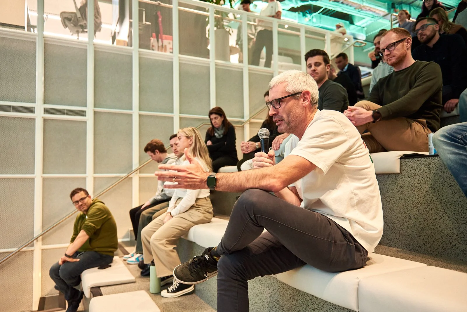 A man with gray hair and glasses sitting on tiered seating, speaking into a microphone during an event or conference, with a diverse group of people seated around him listening attentively.