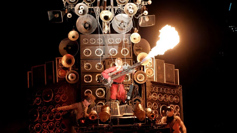 Person performing with a flame-blowing device in front of a wall of large speakers as two other performers help.