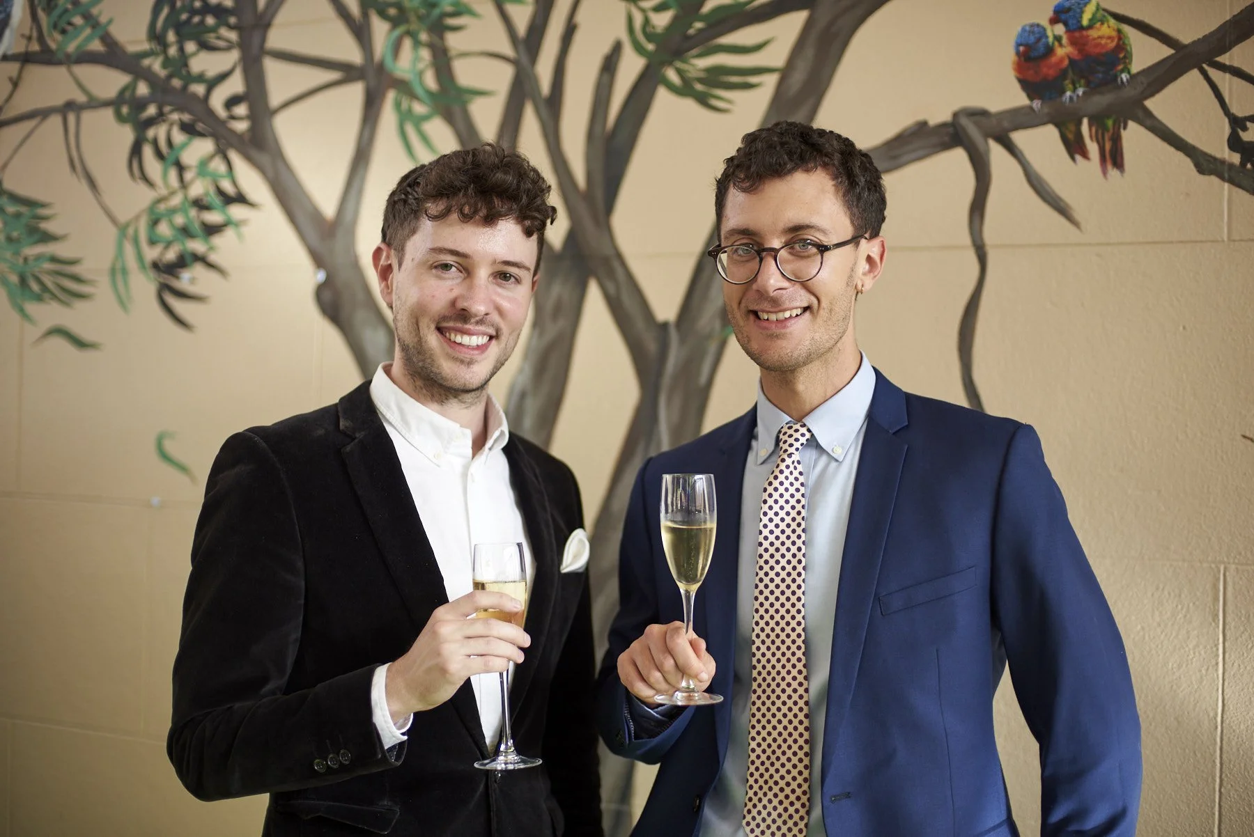 Two men in suits smiling and holding glasses of champagne at a social event with a decorative tree in the background