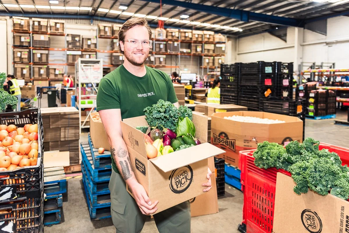 A man in a green Farmers Pick T-shirt holding a box of fresh vegetables, standing inside a warehouse with shelves of items and other workers in the background.