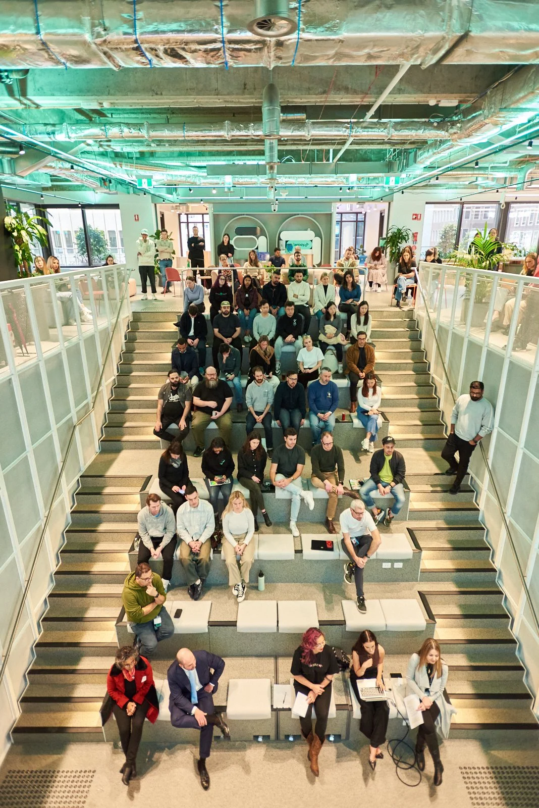 Audience members attending a conference or presentation in a modern, brightly lit, multi-level indoor space with exposed ceiling pipes and large windows.