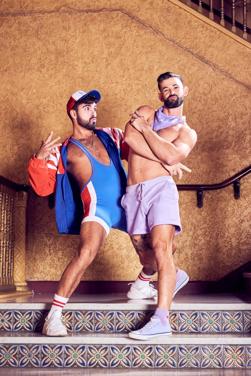 Two young men dressed in sporty, retro-style clothing posing playfully on indoor stairs with a textured beige wall in the background.