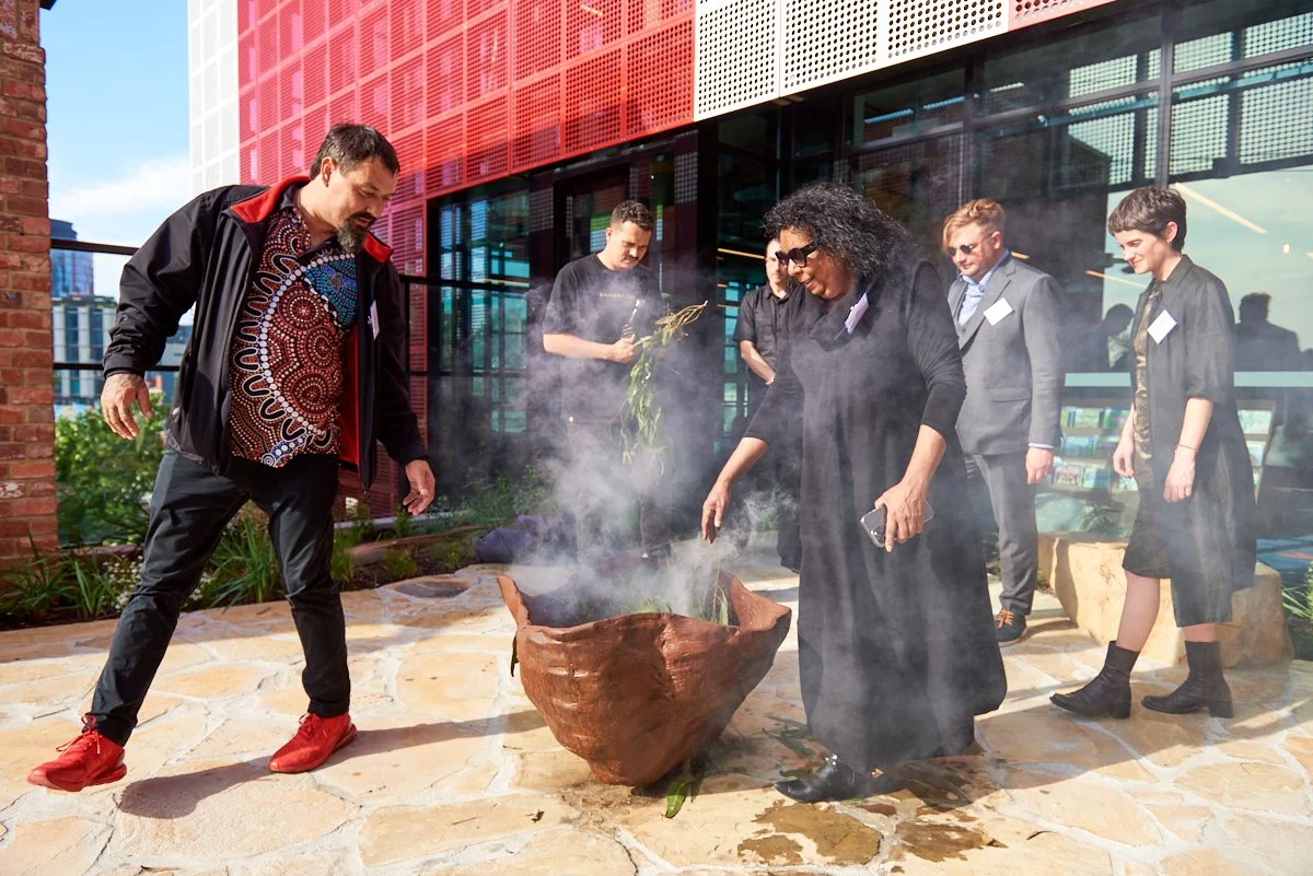 A group of six people gathered outdoors around a steaming, large clay bowl, with some holding items like plants and their phones. They are dressed in a mix of casual and business attire, with a modern building with red and glass panels in the backgro