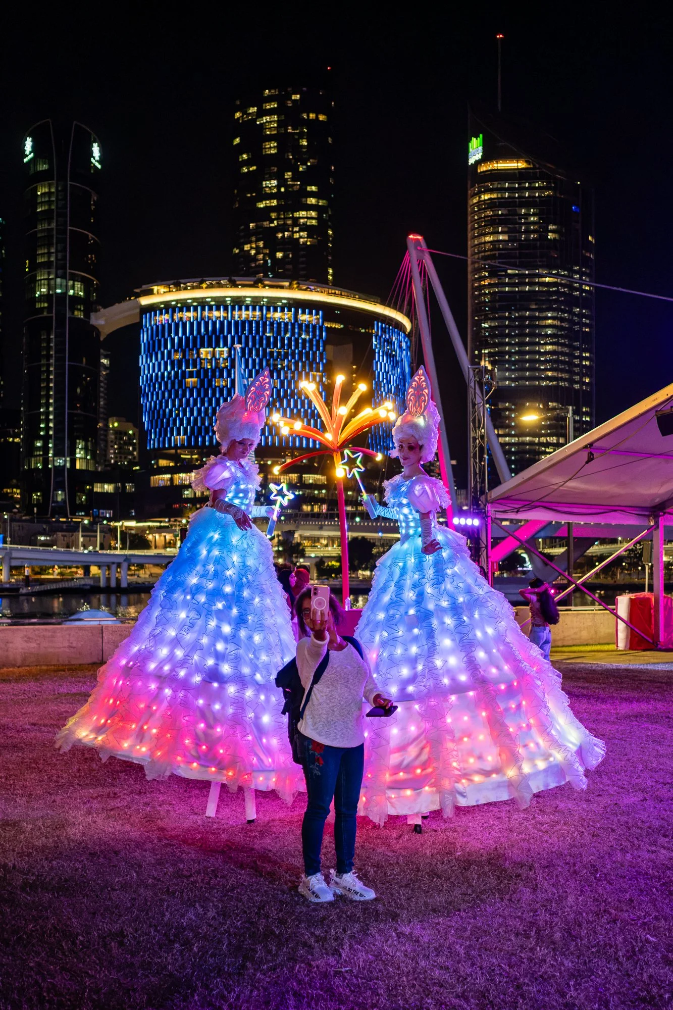 Night scene of two performers in illuminated white gowns with LED lights, standing in front of a city skyline with tall, lit modern buildings and a Ferris wheel with colorful lights.