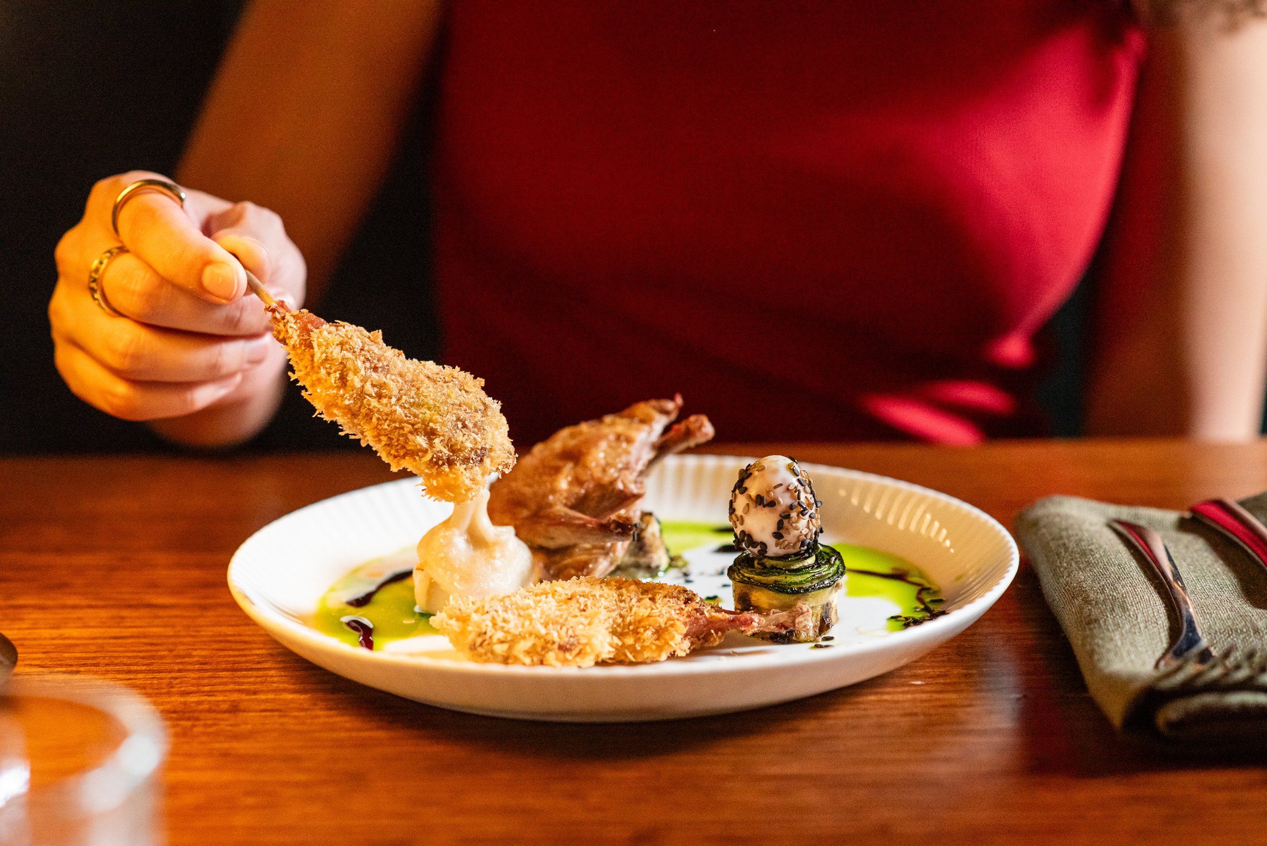 Close-up of a person's hand holding a fried chicken tender over a plate with various gourmet food items, including mashed potatoes, a stuffed mushroom, and sliced meat, on a wooden table.