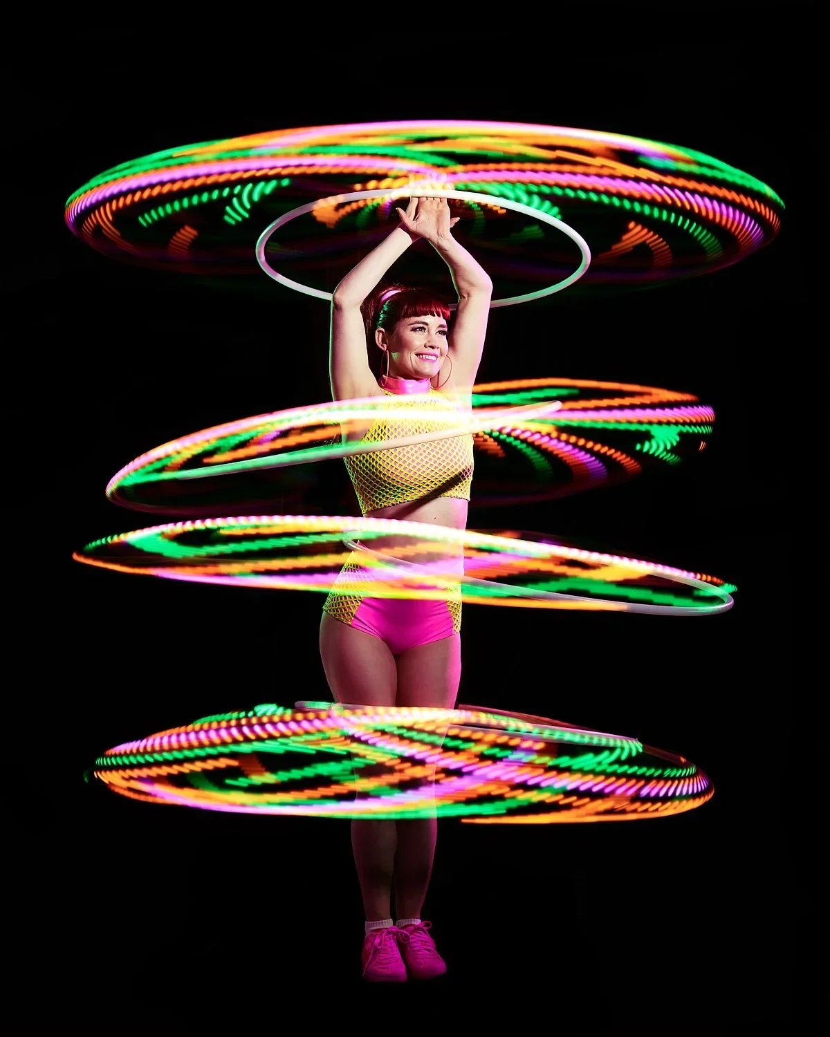 A woman in colorful workout attire spinning LED hula hoops around her body against a black background.