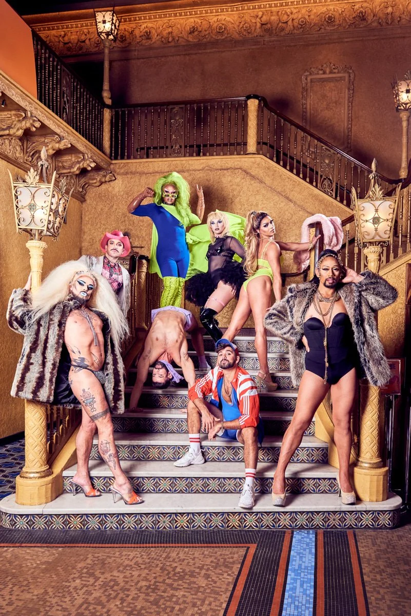 Group of diverse drag performers posing on a staircase in an ornate, vintage-style interior with patterned carpet and warm-toned walls.
