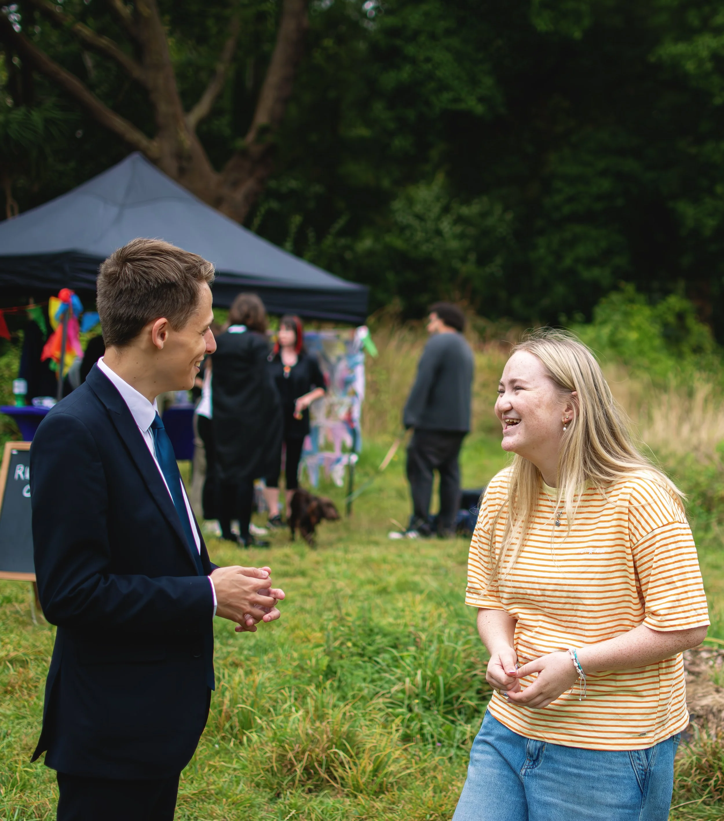 A man in a suit and a woman in a striped shirt are talking and smiling outdoors at a gathering, with a black canopy, people, and trees in the background.