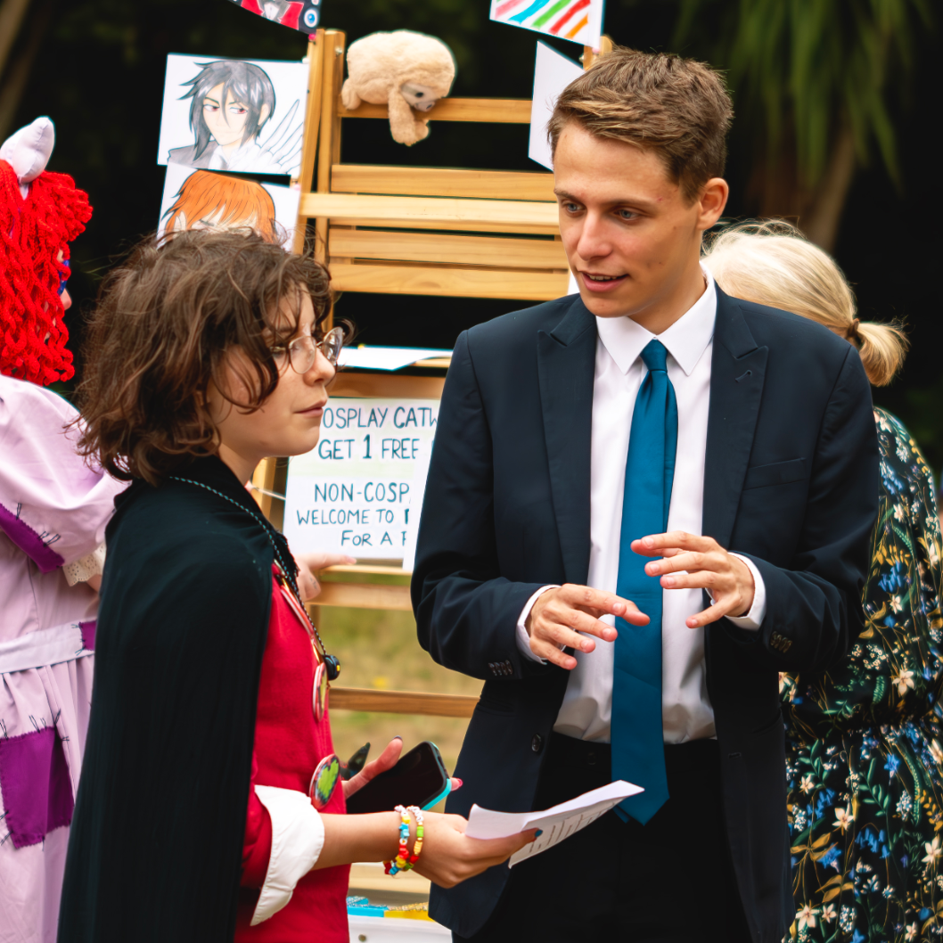 A young boy dressed as a superhero with glasses and a cape talking to a man in a suit at an outdoor event with cosplay and anime drawings in the background.