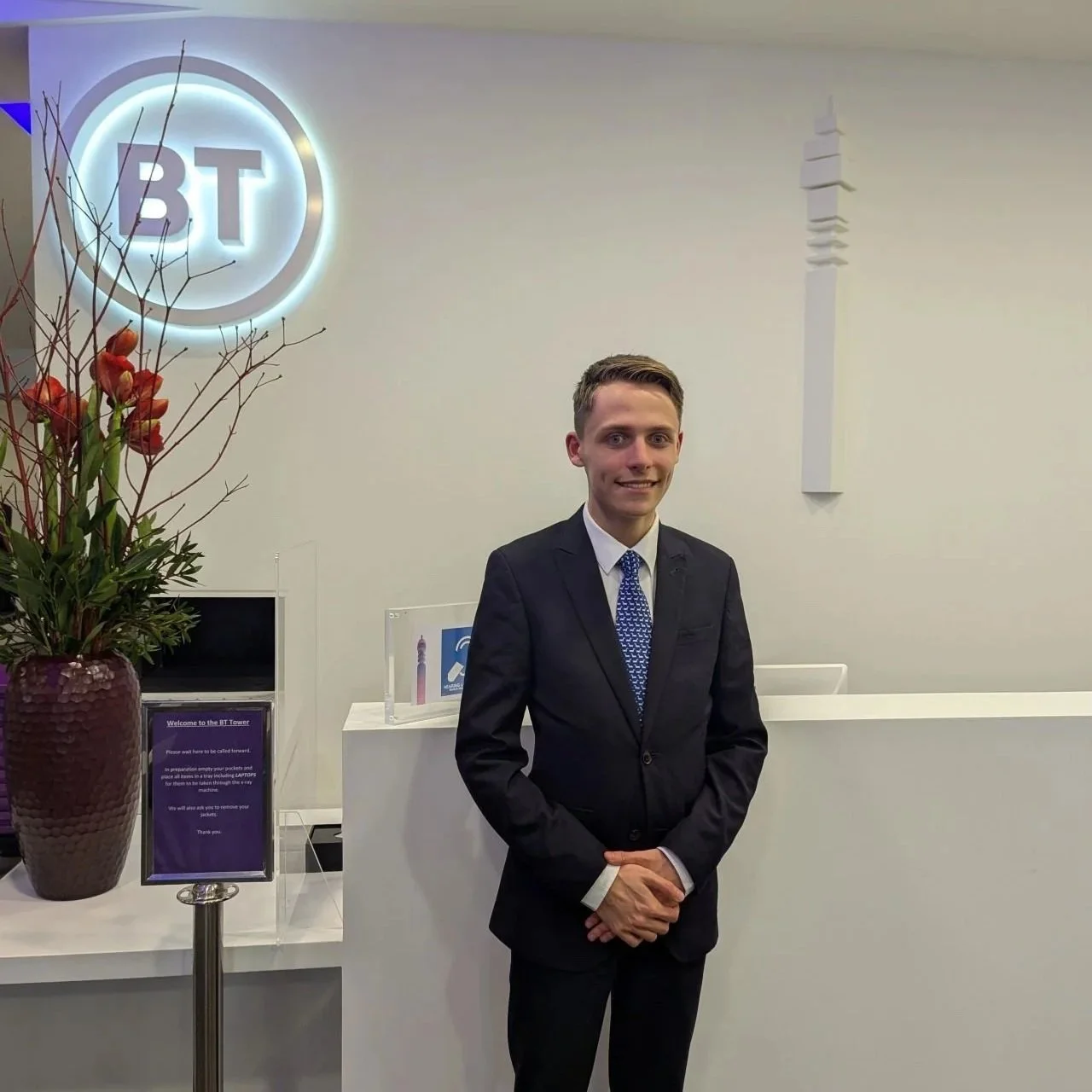 A young man (Kole Gjikolaj) in a black suit and blue tie standing in front of a white reception desk at the BT Tower, with a large purple flower arrangement on his left and a purple sign.