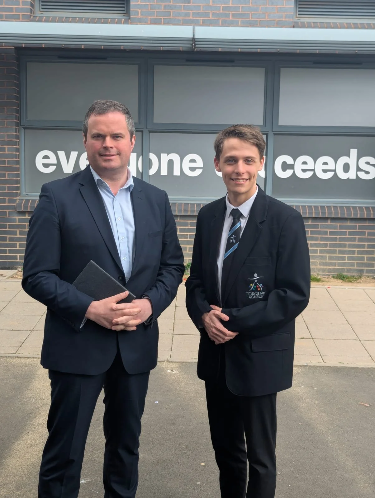 Kole Gjikolaj and Kevin Foster standing outside in front of a window with large text that reads 'everyone succeeds'. One man is holding a black folder and the other has a crest on his blazer that reads 'Torquay Academy'.