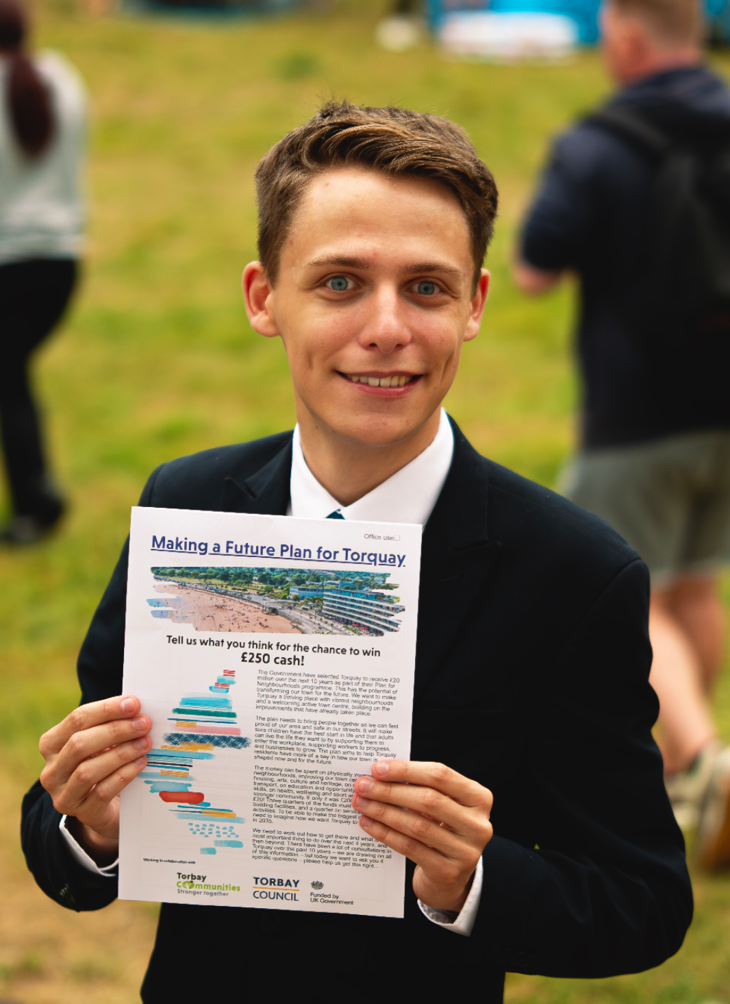 A young man in a black suit and tie holding a flyer about making a future plan for Torquay, promoting a chance to win £250 cash, at an outdoor event with grass and blurred people in the background.