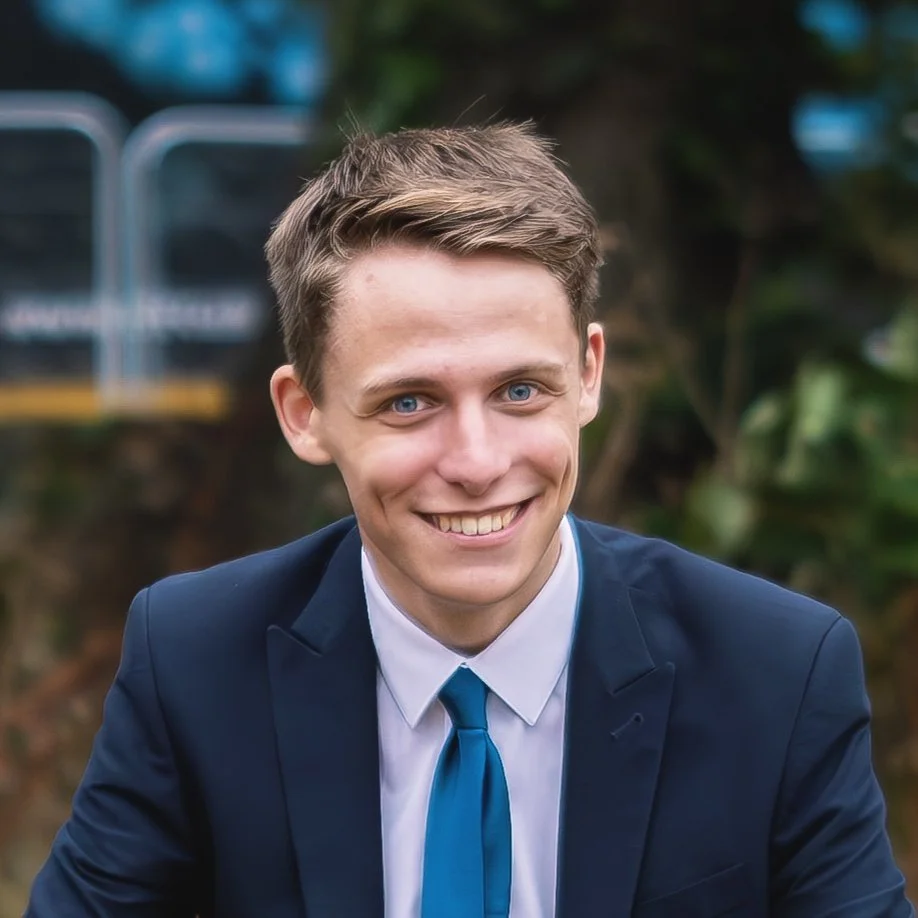 A young man with light skin, blue eyes, and short brown hair in a suit and tie smiling outdoors.