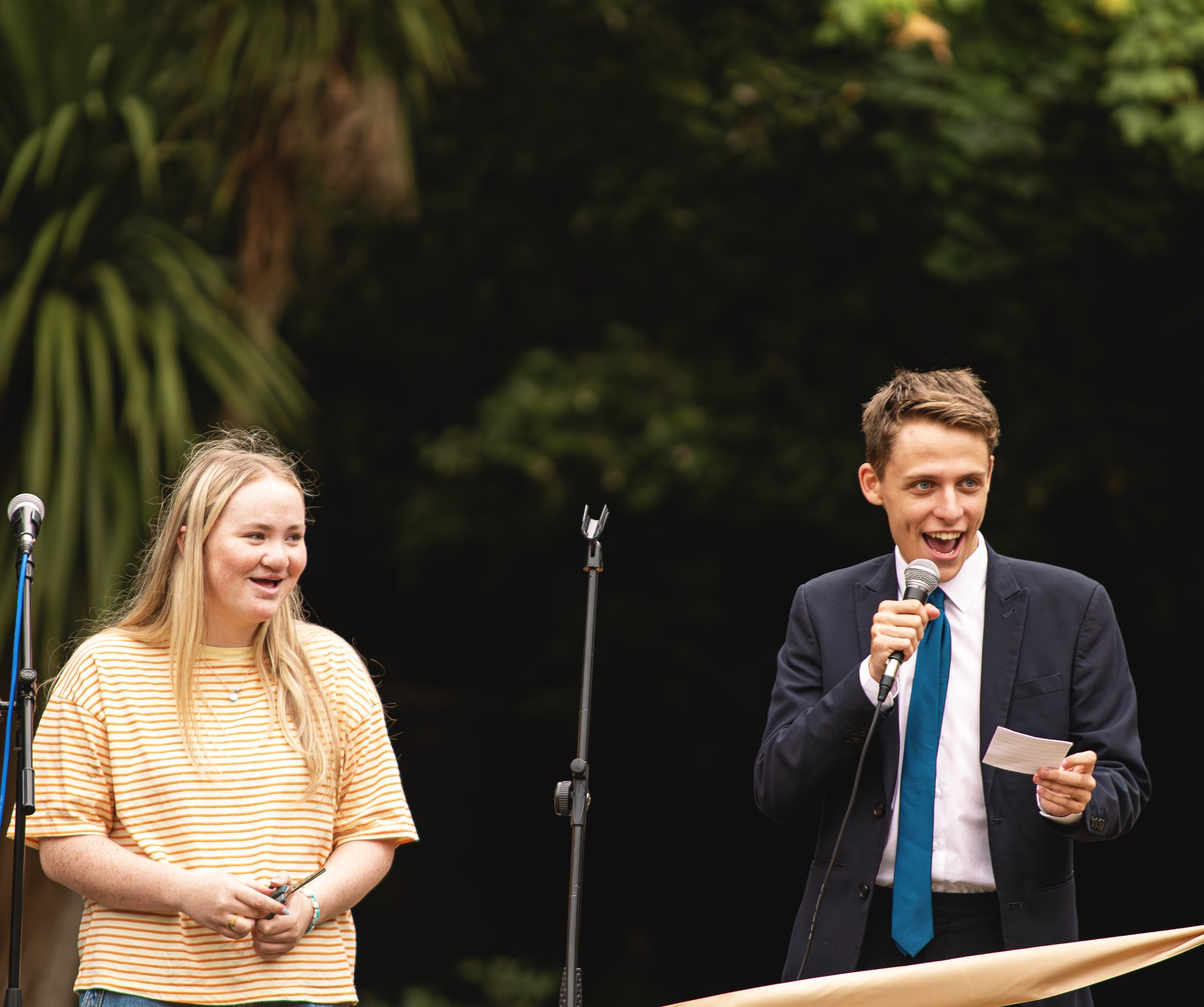 Two young individuals speaking into microphones outdoors, one woman in a yellow striped shirt and a young man (Kole Gjikolaj) in a suit with a blue tie, smiling.