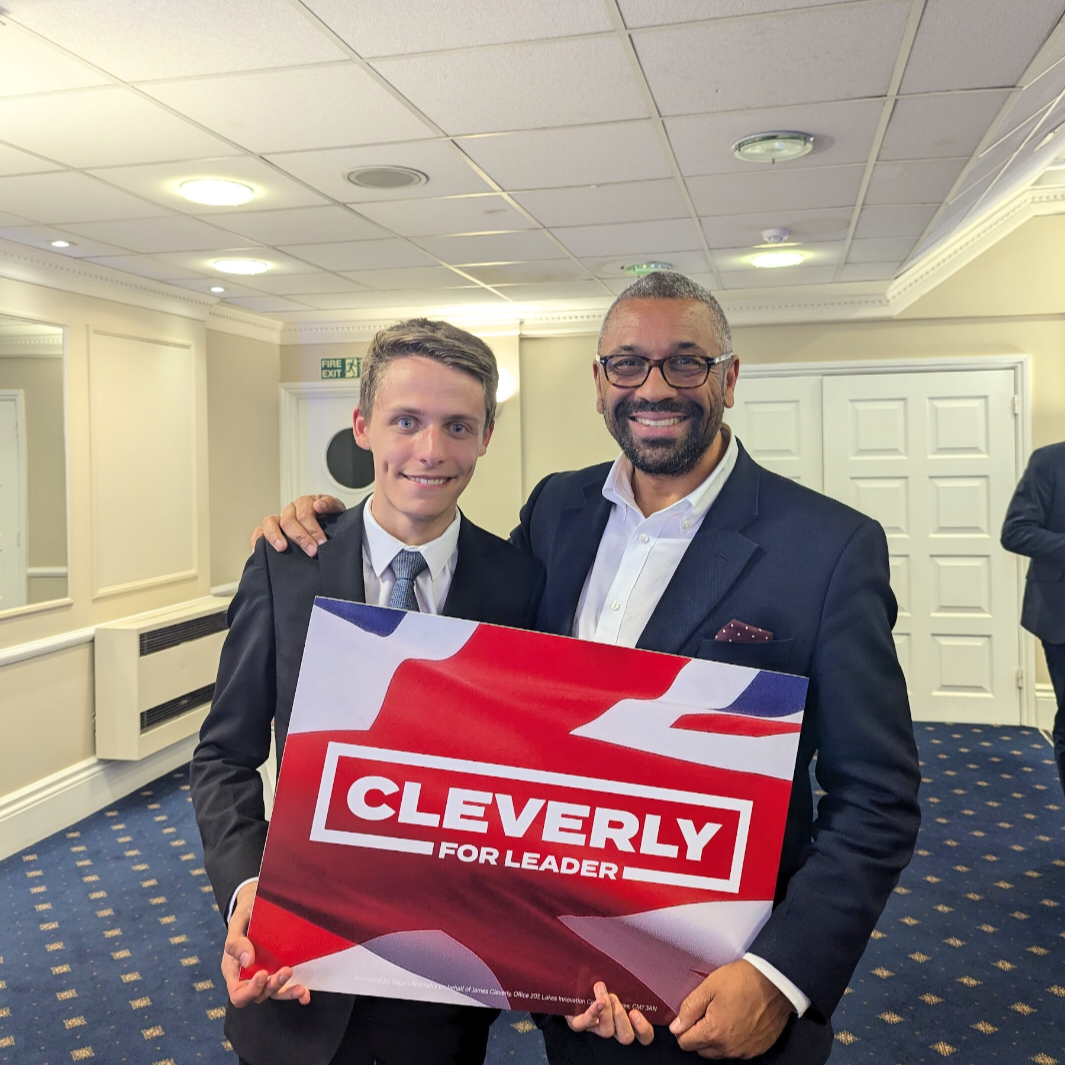 Two men in suits smiling, holding a campaign sign that says 'Cleverly for Leader,' in a well-lit room with white walls and blue carpet.