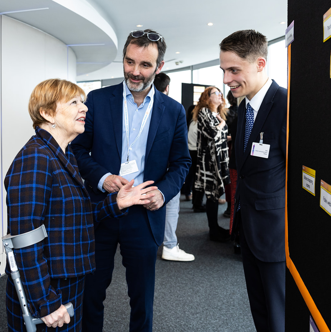 Three people engaged in conversation at a professional event or conference. An elderly woman using a cane and wearing a plaid blazer talks with a man and a younger man in suits, with name badges, in a modern conference room with other attendees in the background.