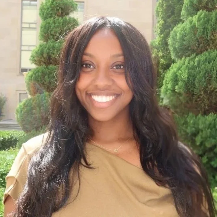 A woman smiling outdoors with green bushes and trees in the background.
