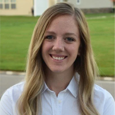 A young woman with long blonde hair smiling outdoors in front of a green lawn and buildings.