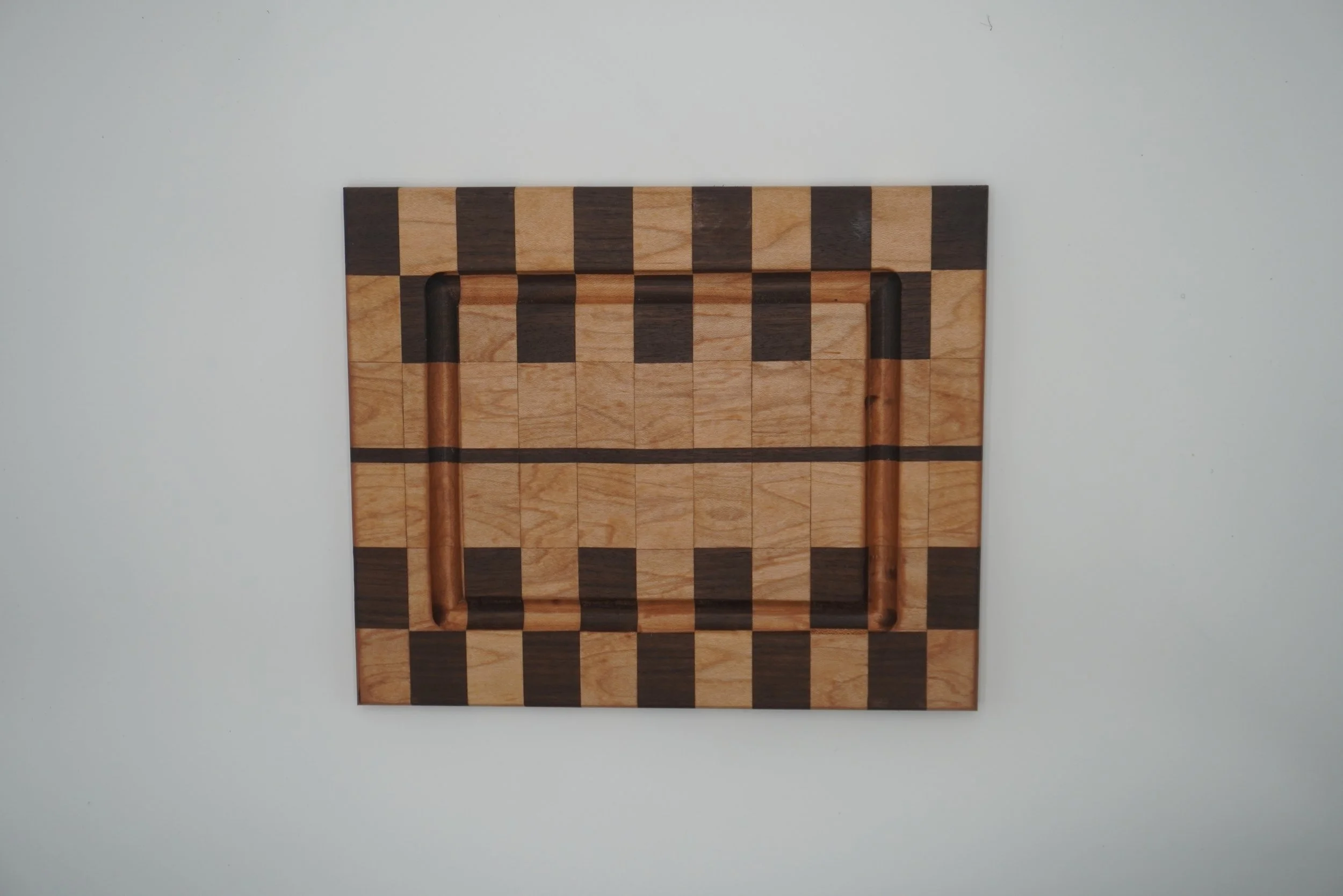 A wooden chessboard with alternating light and dark squares, featuring cutouts for chess pieces, mounted on a plain white wall.