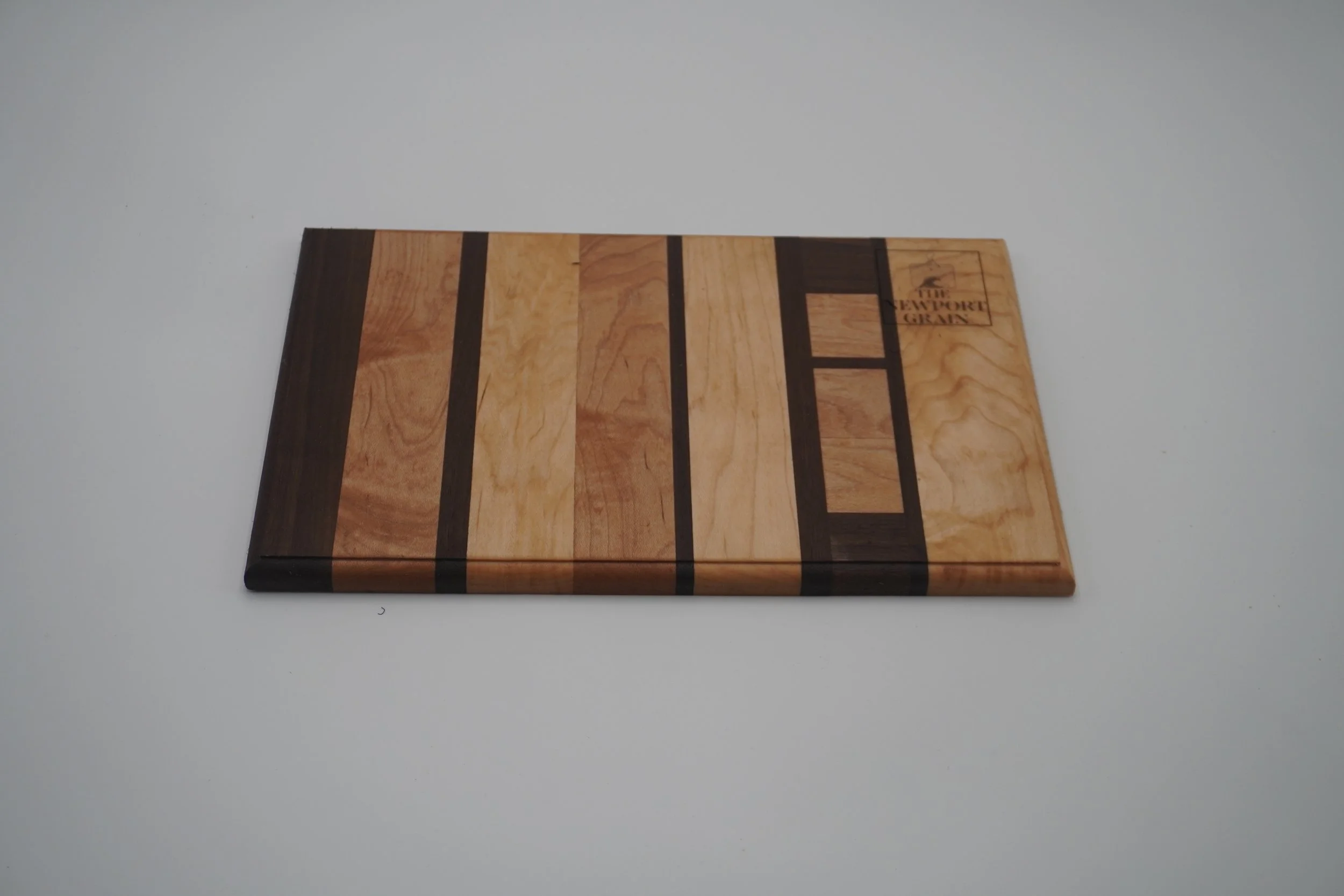 A rectangular wooden cutting board with a striped pattern of light and dark wood, featuring the engraved logo of The Newport Grain in the top right corner.