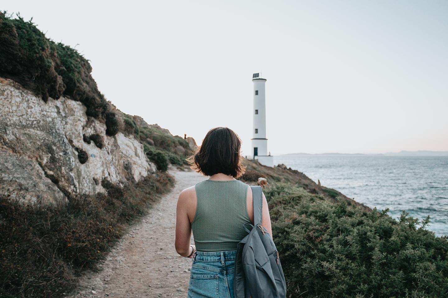 Woman with short dark hair, wearing a sleeveless green top and jeans, carrying a gray backpack, walking along a dirt path near the coast with a lighthouse in the background.