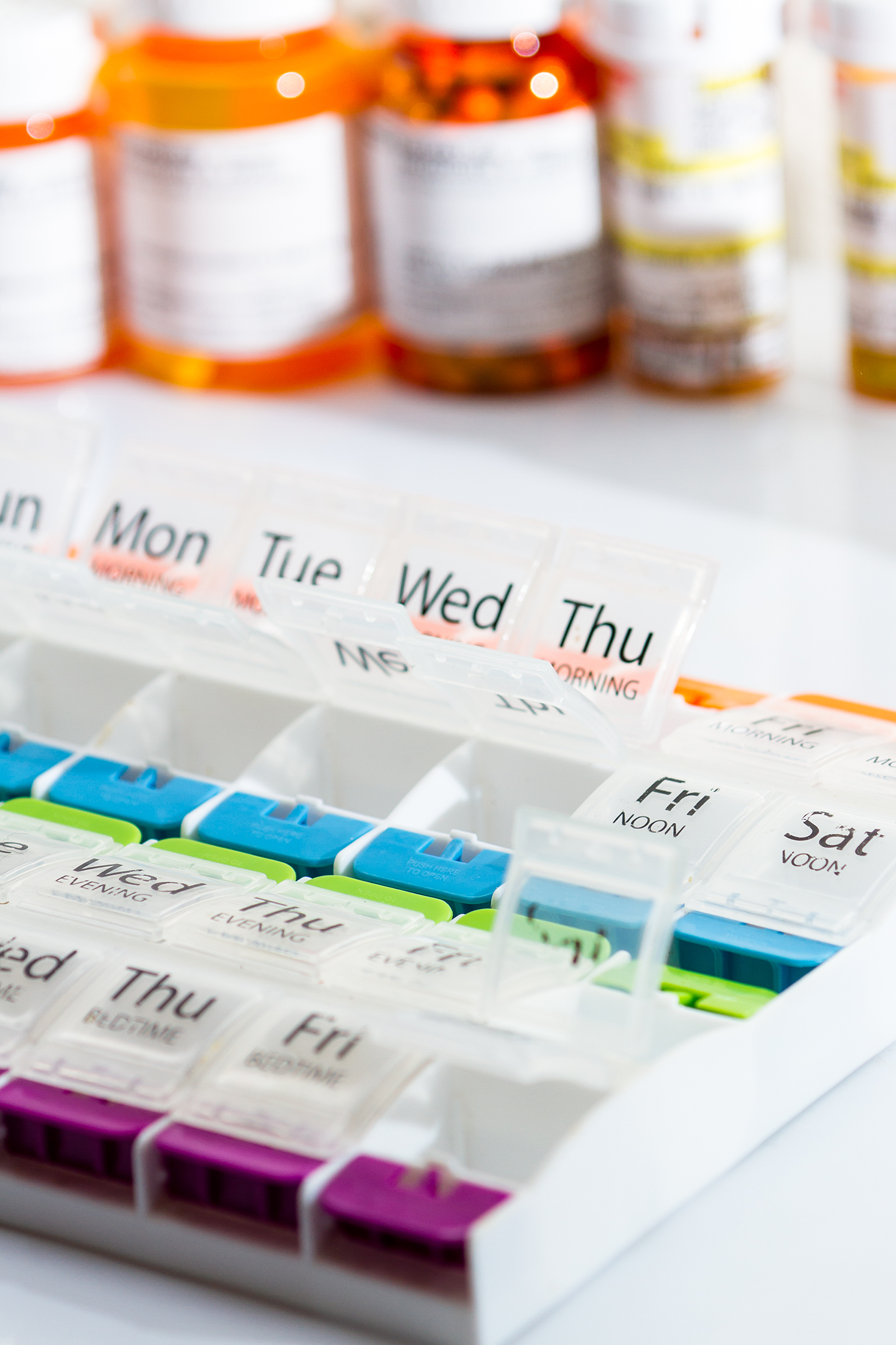 Pill organizer with days of the week labeled, filled with medication for each day, and prescription pill bottles in the background.