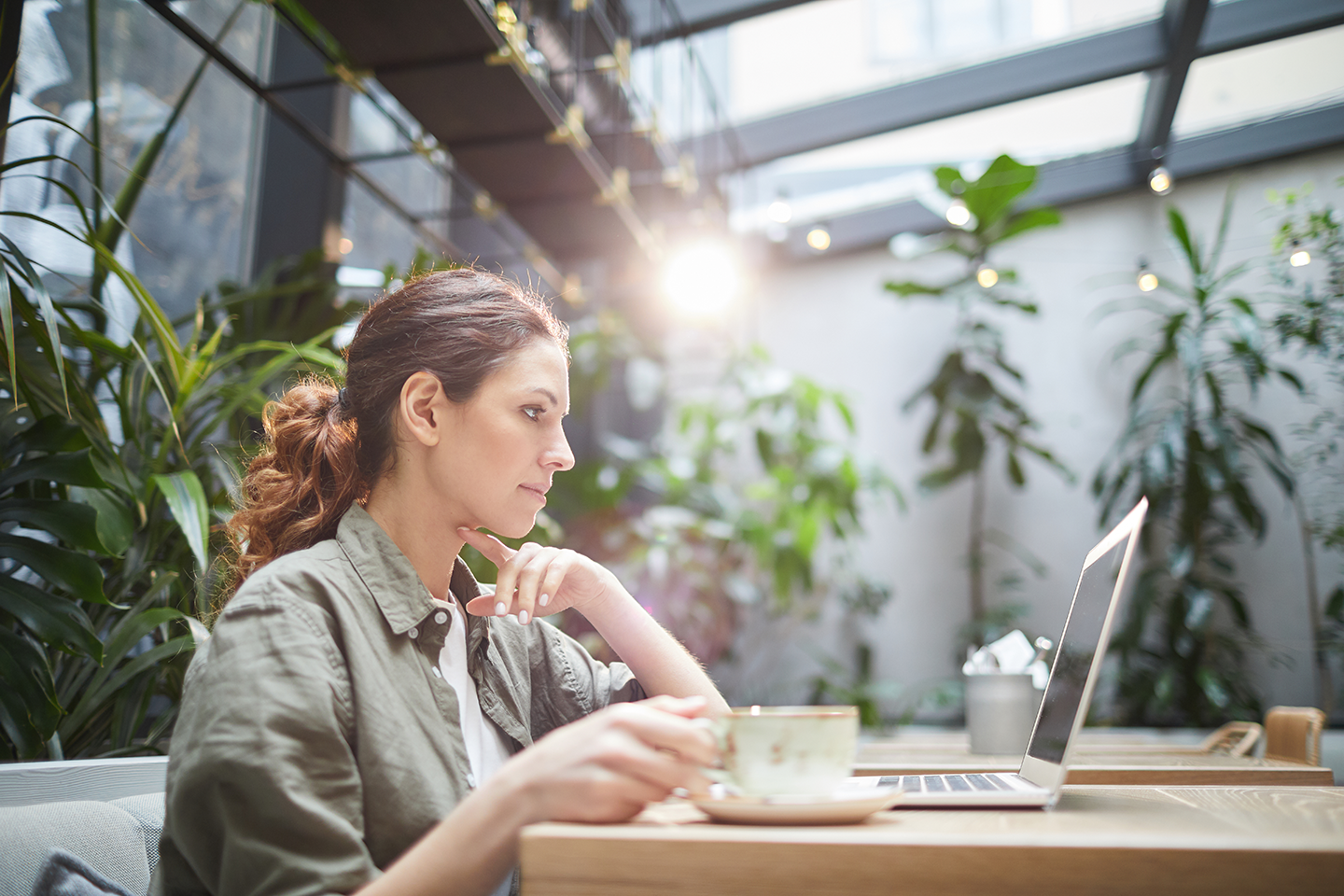 Woman working on a laptop in a bright greenhouse with green plants and sunlight.
