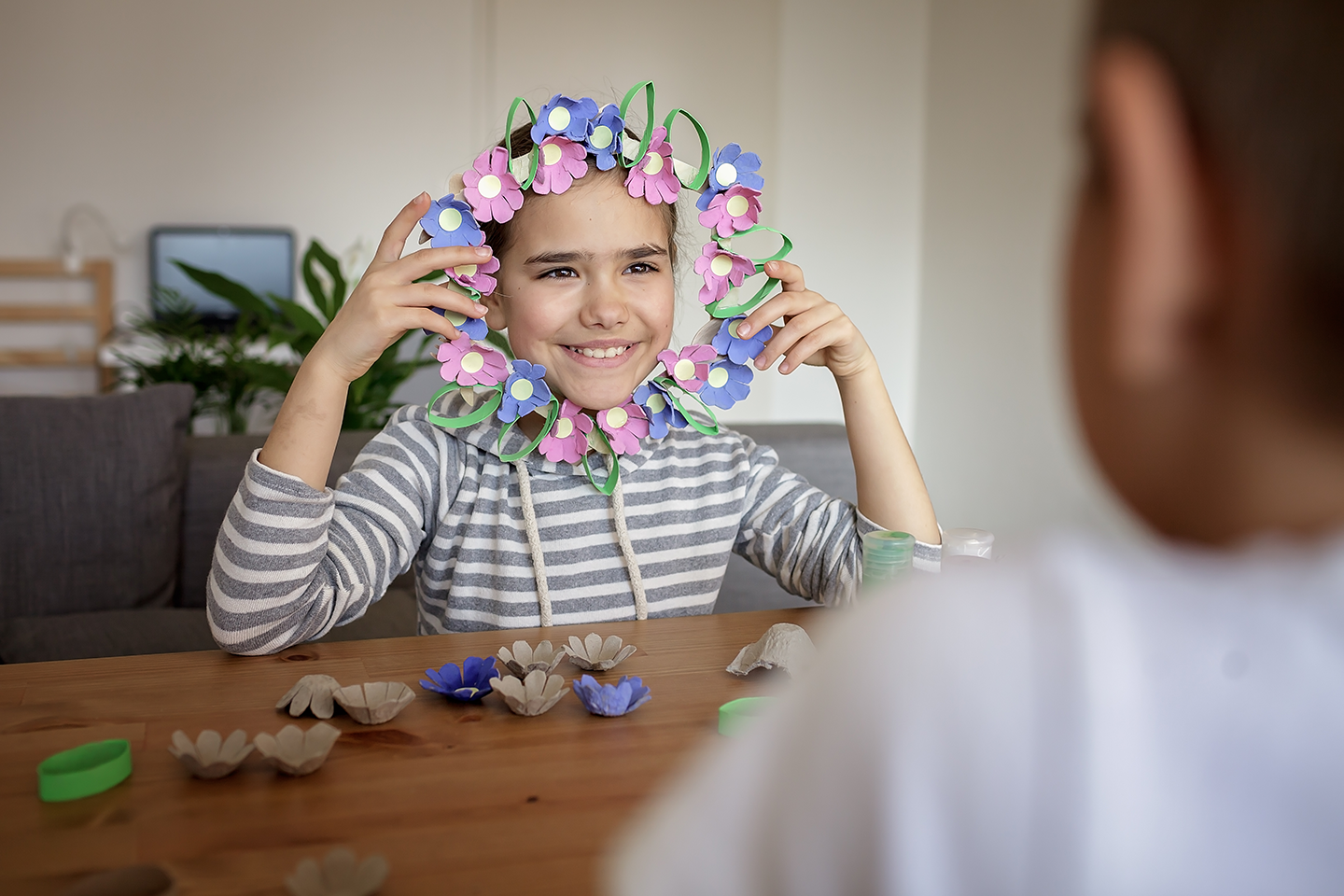Girl wearing a paper flower wreath, smiling, and sitting at a table with paper flowers and craft supplies.