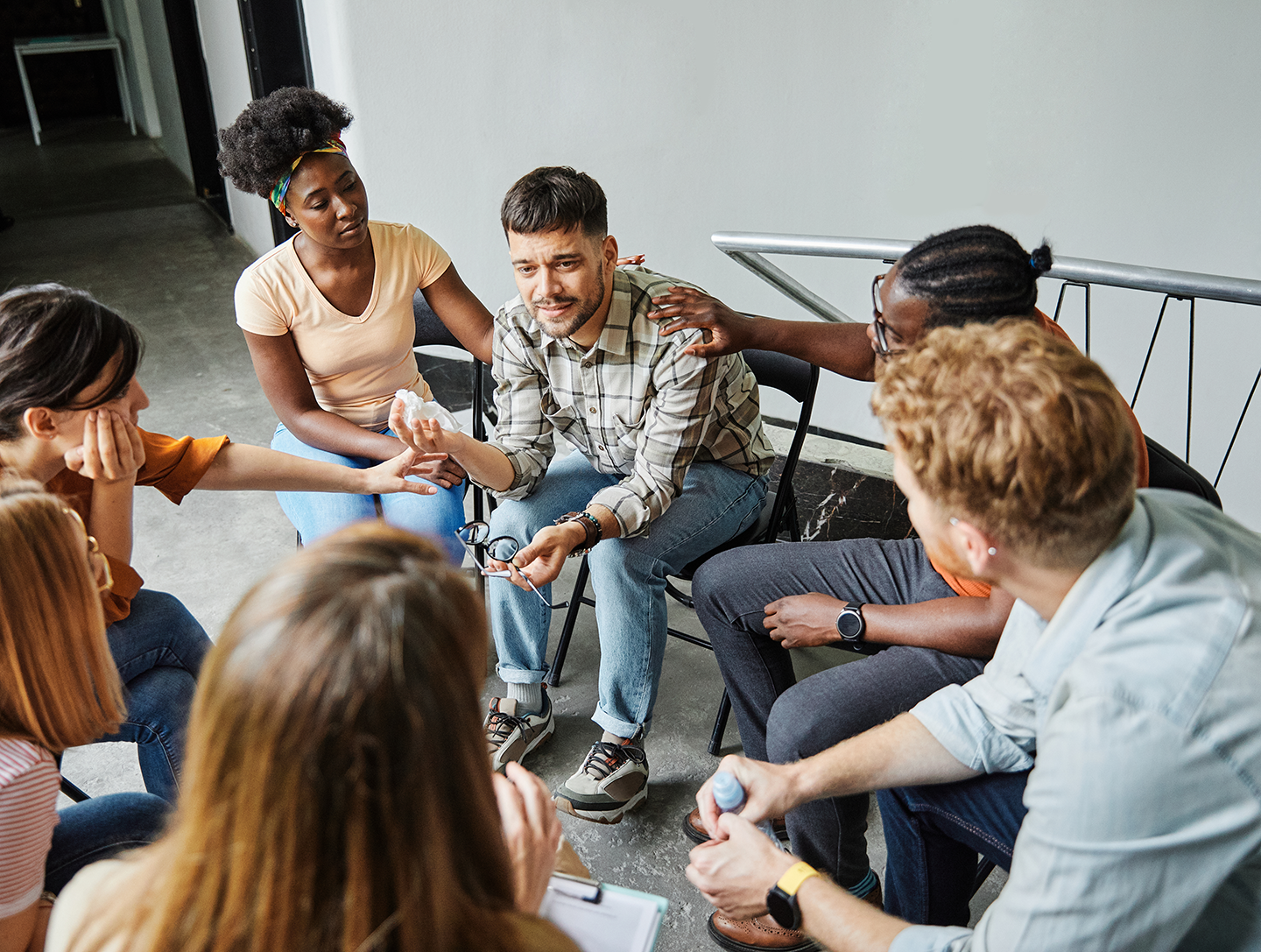 A group of diverse people sitting in a circle, comforting a young man who appears upset, in a support group setting.