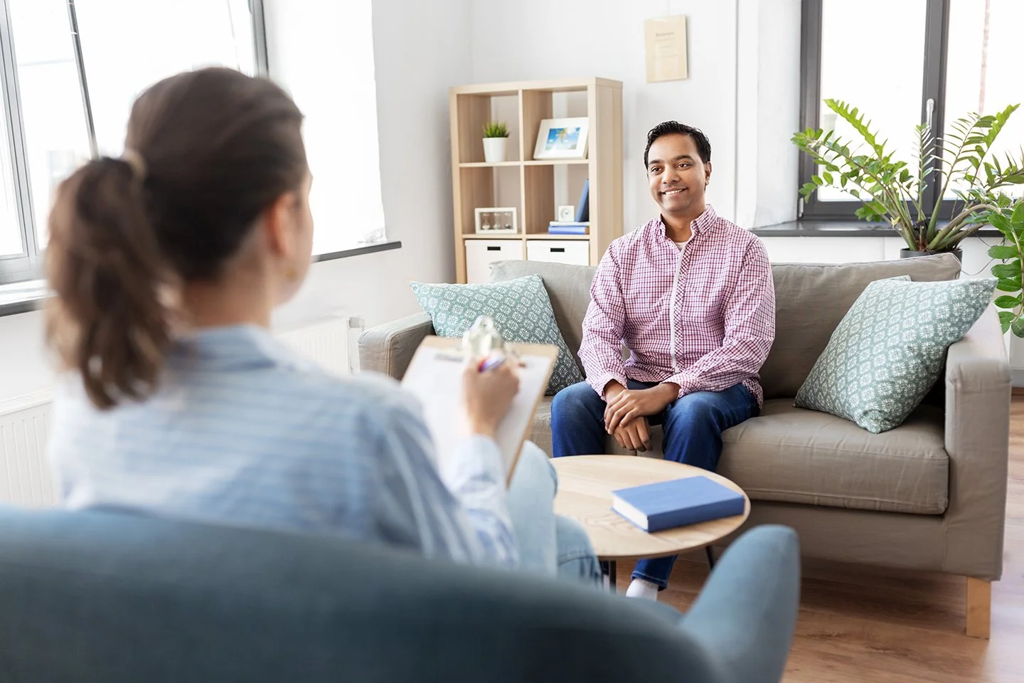 A man sitting on a beige sofa smiling during a therapy or counseling session with a female therapist taking notes on a notepad in a bright, modern office.