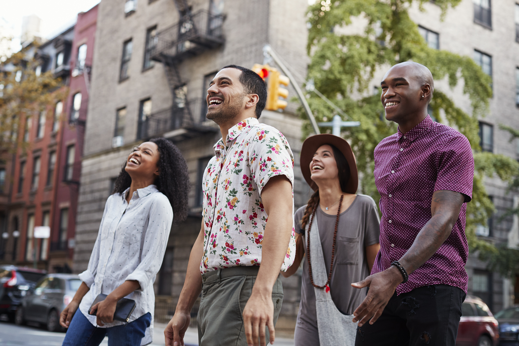 Four diverse young adults walking on city street, smiling and enjoying themselves, with residential buildings and a traffic light in the background.