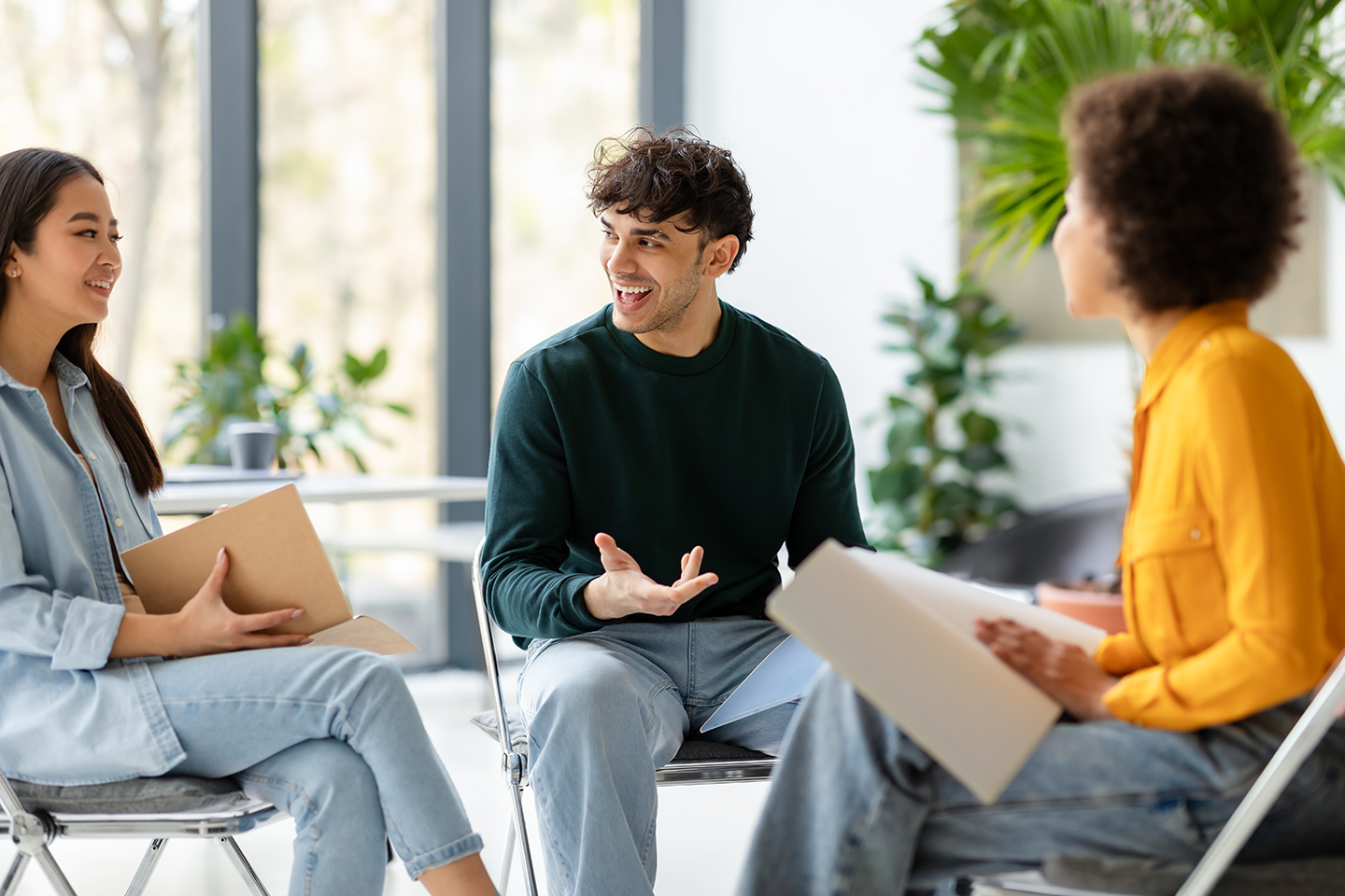 Four people having a discussion in a bright room with large windows and green plants.