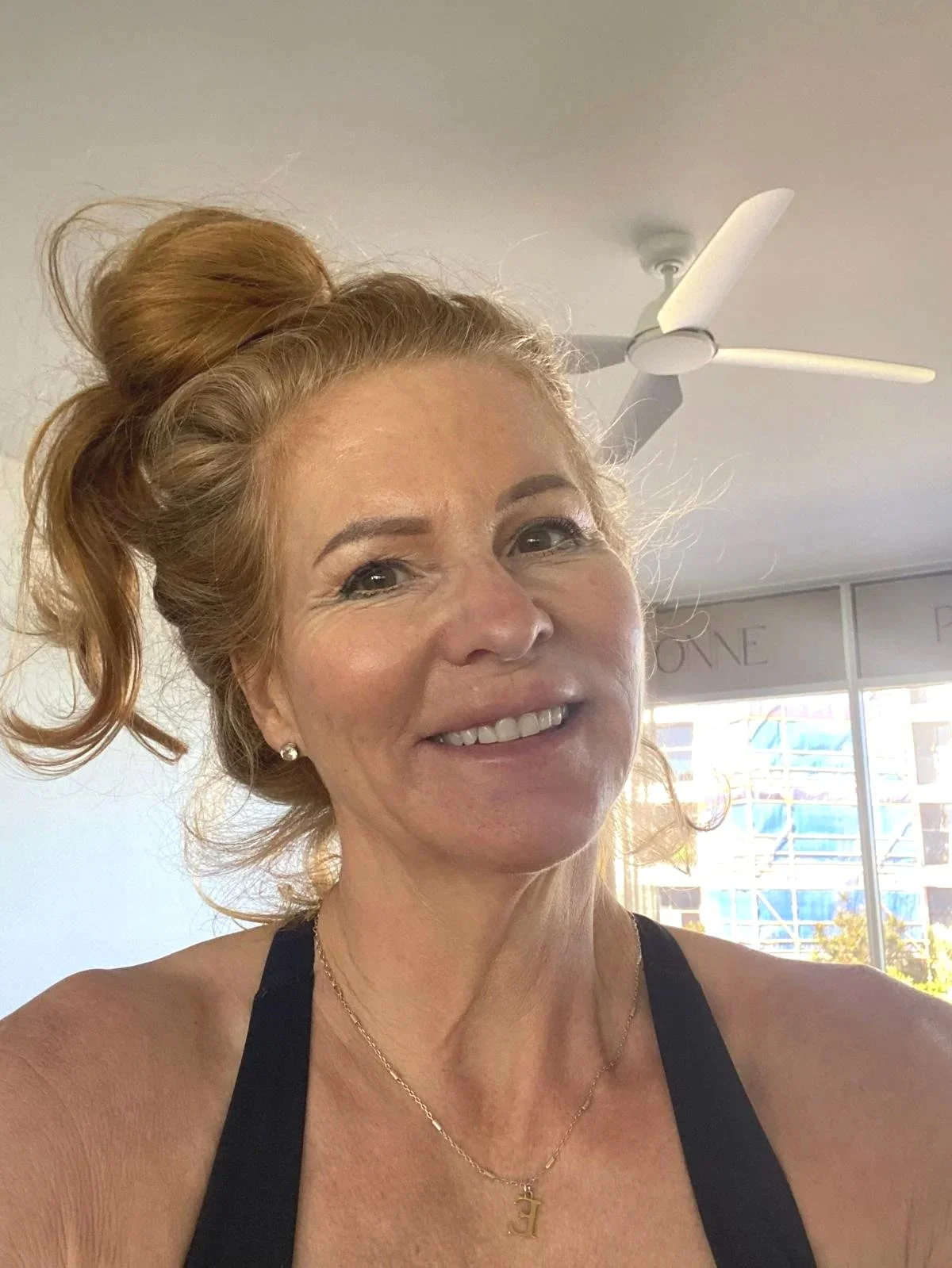 A woman with red hair tied in a bun, smiling, wearing a black sleeveless top, earrings, a gold necklace, and a pendant.