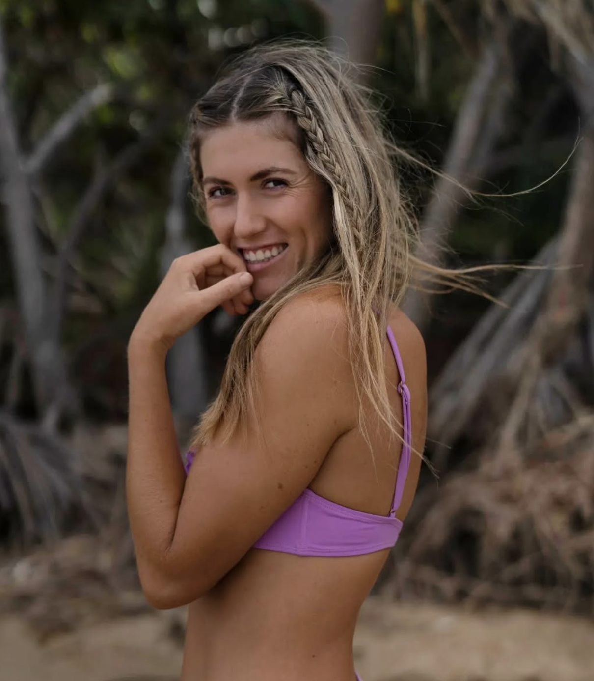 A smiling woman with long, wavy blonde hair with a small braid, wearing a purple bikini top, standing outdoors with trees and branches in the background.