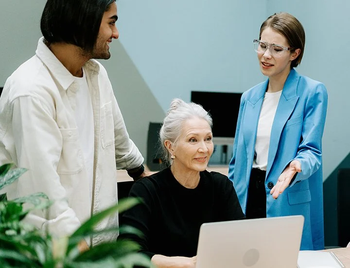 Three people having a discussion in an office. An elderly woman is seated at a desk with a laptop. A man and a woman are standing beside her, engaging in conversation.