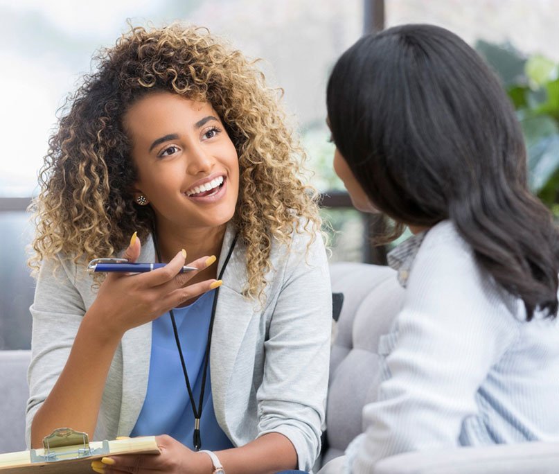 Two women having a conversation, one with curly hair and a stethoscope, appearing to be a healthcare professional, smiling and gesturing with a pen.