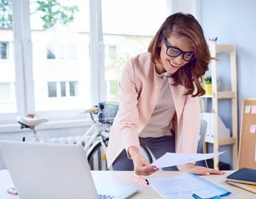 A woman in glasses and a pink blazer looks at papers on a desk in a bright office with large windows and a bicycle in the background.