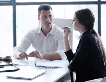 Two people in a professional setting having a conversation, with documents on the table and a large window in the background.