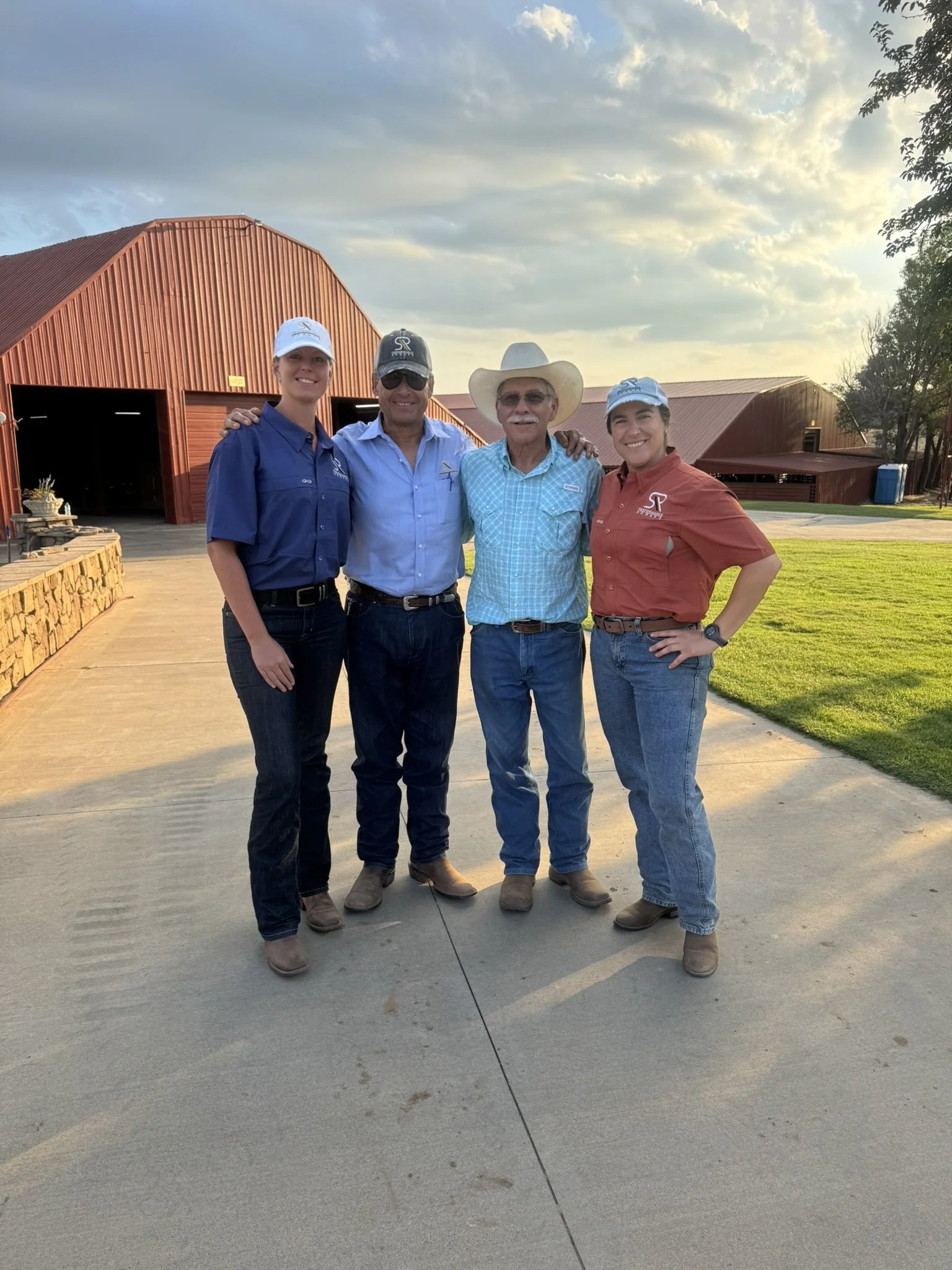 Four individuals standing together outdoors in front of a red barn, smiling, during sunset. Two women on each end and two men in the middle, wearing casual clothes and hats. SR Bar Ranch Performance Horses Boerne Texas USA