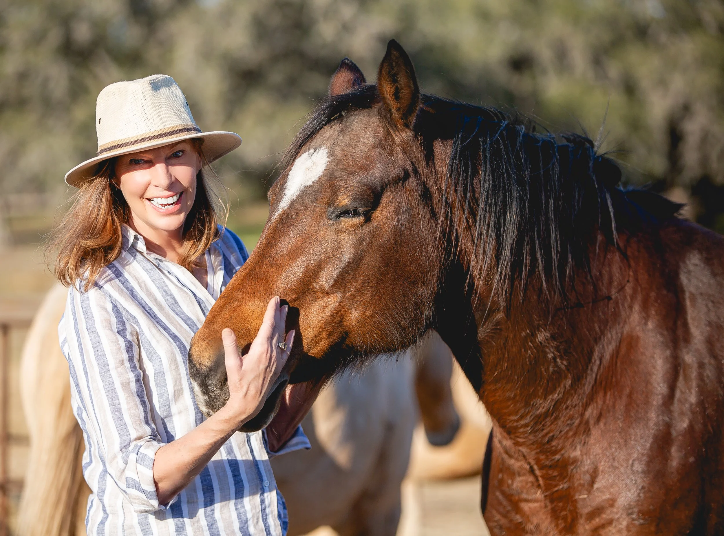 A woman with a hat and striped shirt is gently touching and smiling at a brown horse outdoors. Teri Secrest. SR Bar Ranch Performance Horses Boerne Texas USA. 