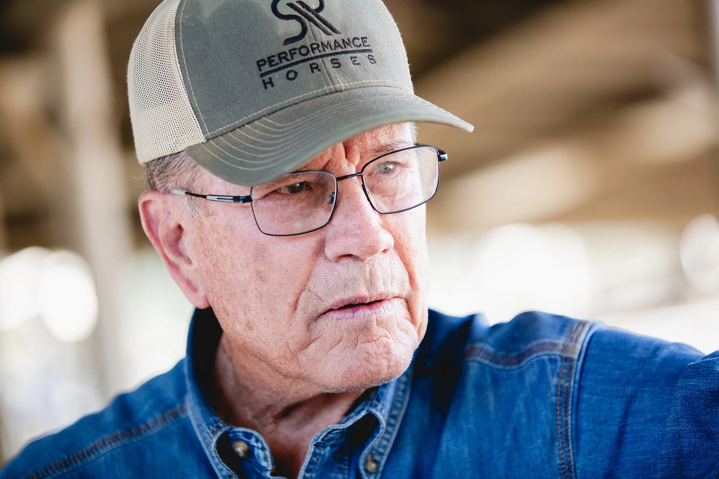 Close-up of an elderly man wearing glasses, a cap with the words "SR Bar Performance Horses," and a denim shirt, in an outdoor setting.