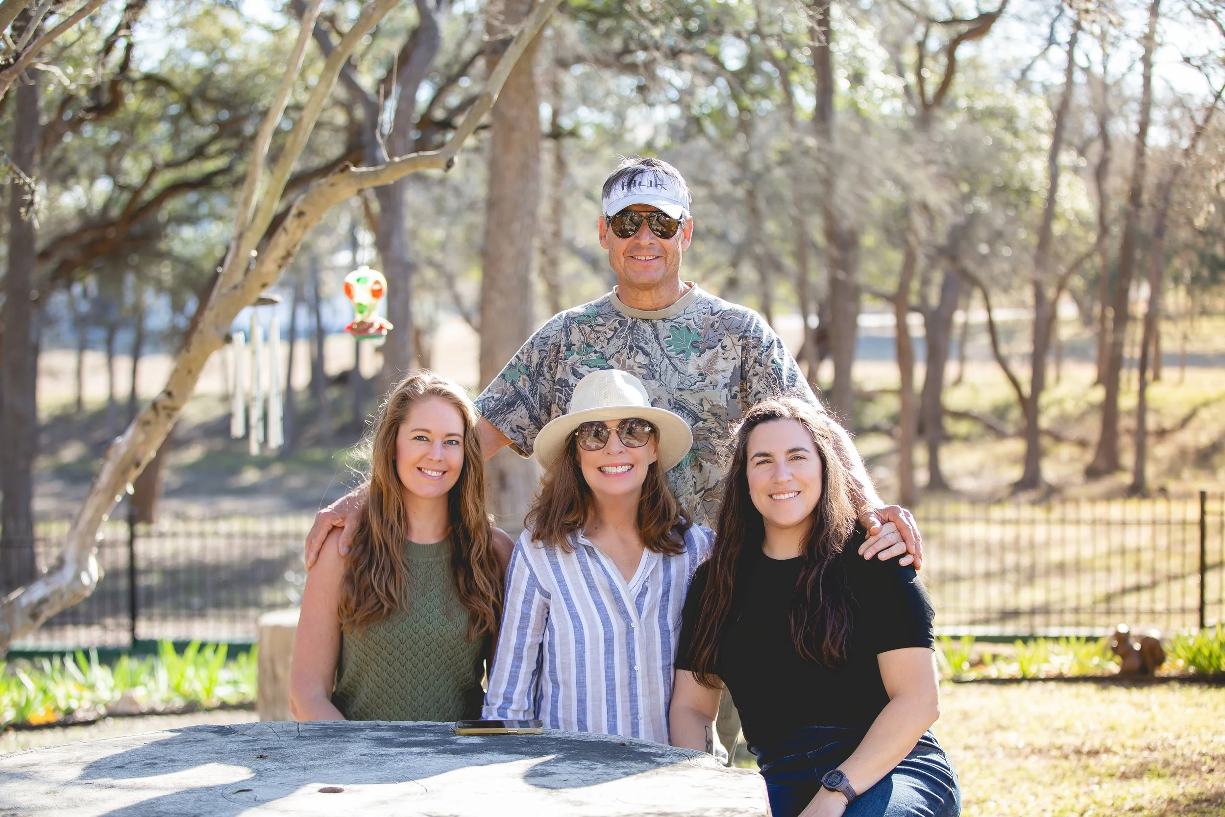 A group of four people smiling outdoors in a park on a sunny day. A man is standing behind three women, with his arms around two of them. The women are sitting at a stone table. All are dressed casually and wearing sunglasses. 